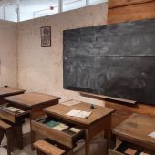 Cztery drewniane ławki szkolne stoją w rzędzie przed tablicą, z lewej strony jest okno