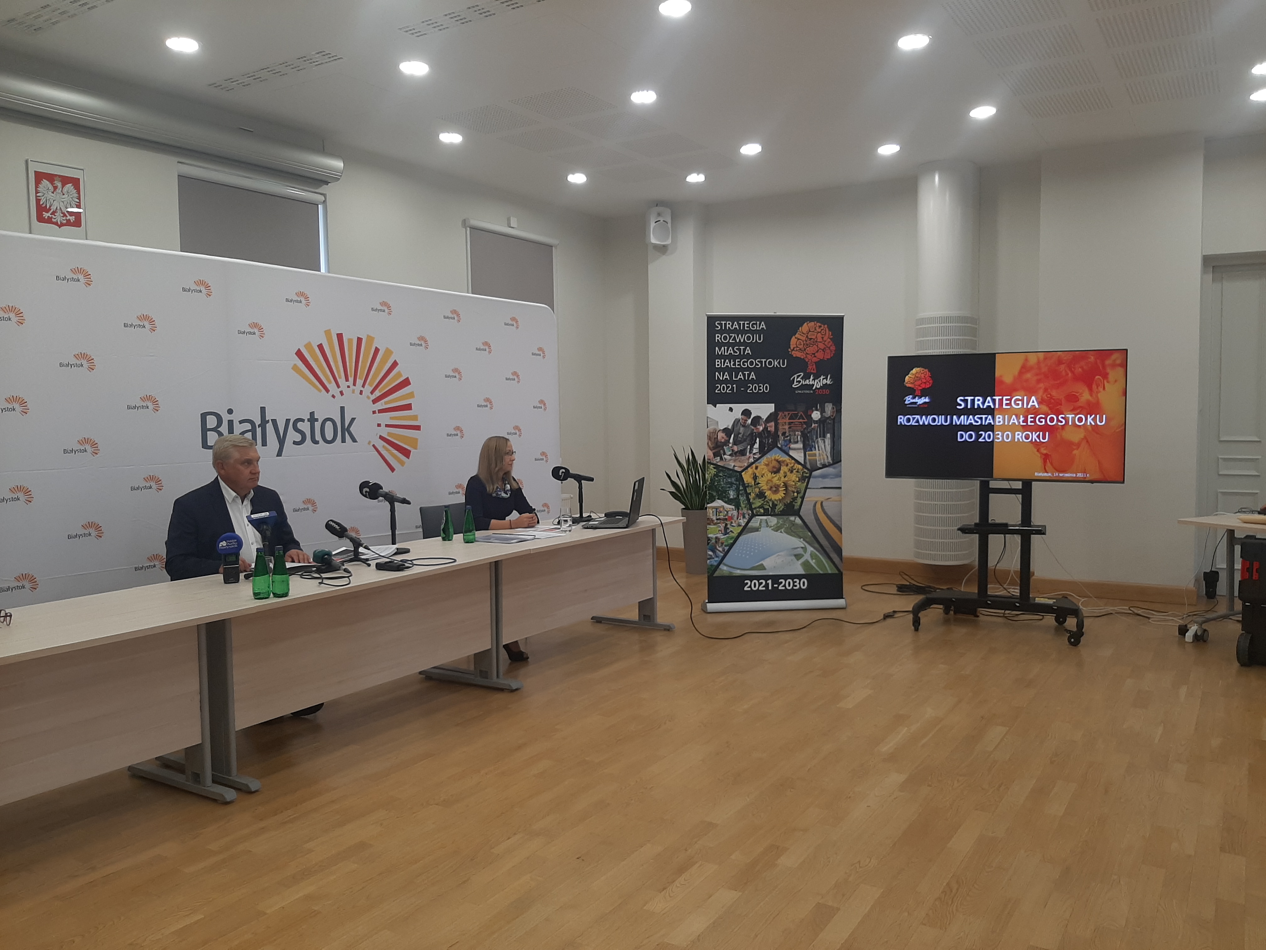 Sala konferencyjna, z lewej strony stoi ścianka z napisem Bialystok, na jej tle przy stole siedzi prezydent Białegostoku oraz kobieta, na wprost stoi stand, lekko na prawo telewizor z wyświetlonym napisem: Strategia Białegostoku