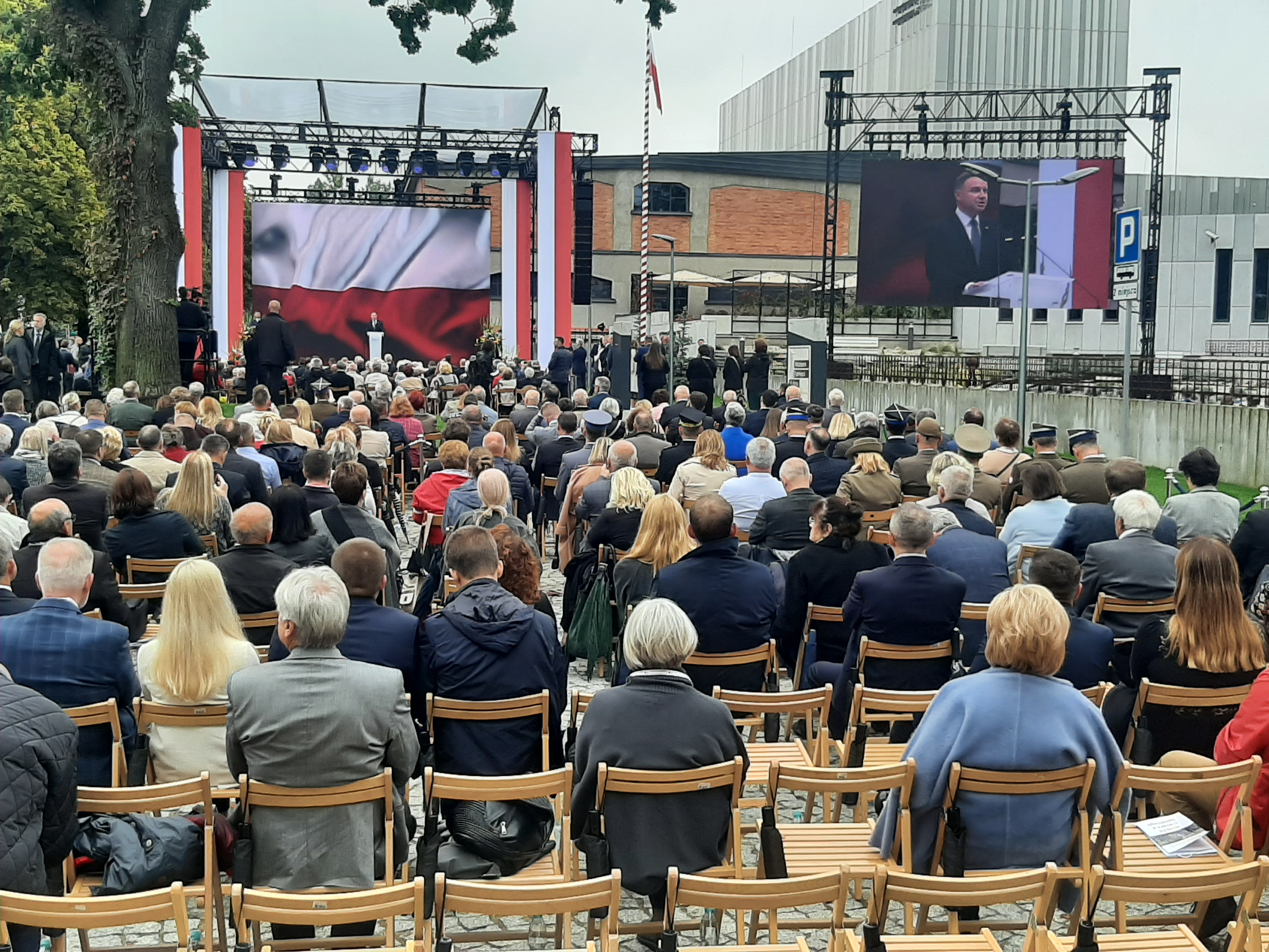 Tłum ludzi siedzi na krzesłach tyłem, a przodem do sceny, na której przemawia prezydent RP Andrzej Duda. Z prawej strony znajduje się telebim, na którym wyświetlona jest postać prezydenta Polski