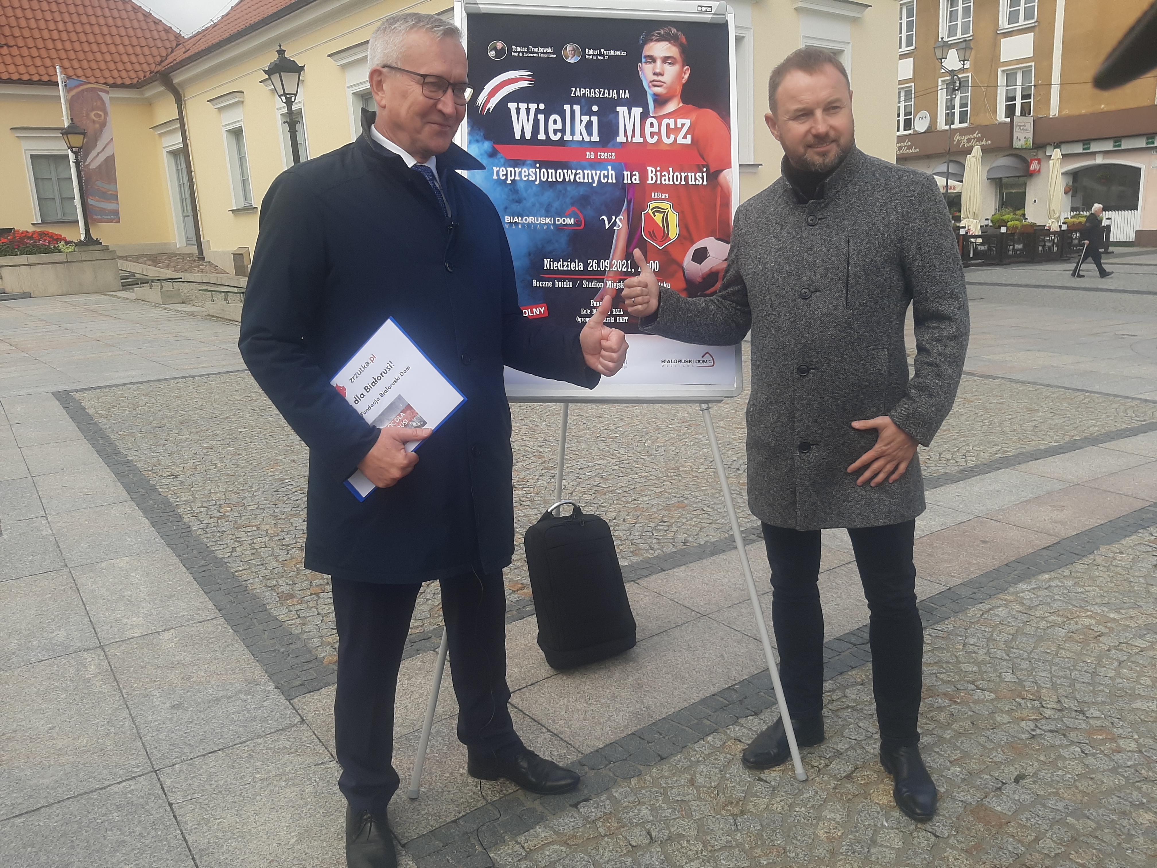 Dwóch mężczyzn stoi przy plakacie promującym charytatywny mecz piłki nożnej