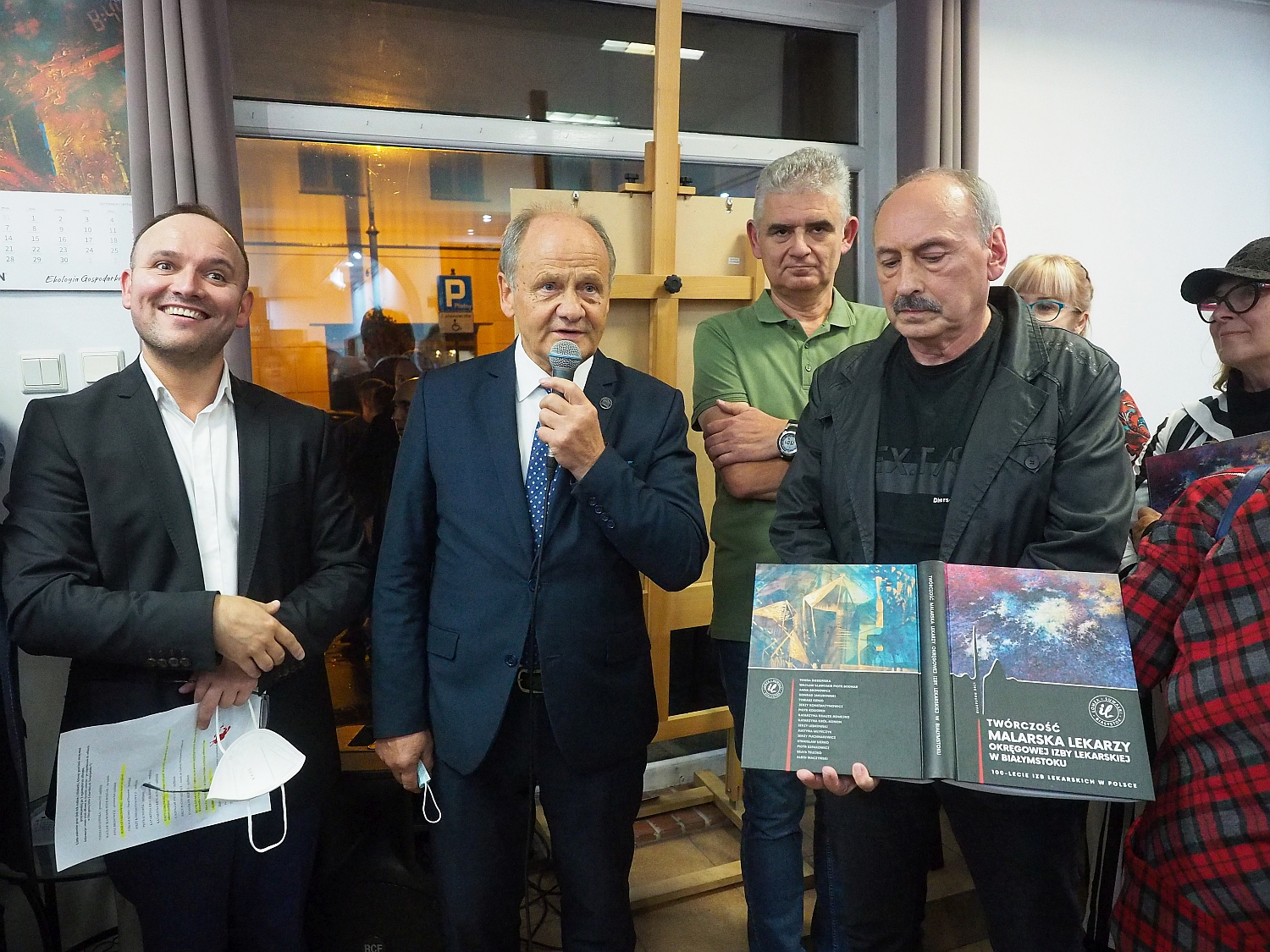 Wernisaż wystawy. Grupka osób stroi obok siebie. Jeden z mężczyznmówi do mikrofonu, drugi prezentuje album fotograficzny