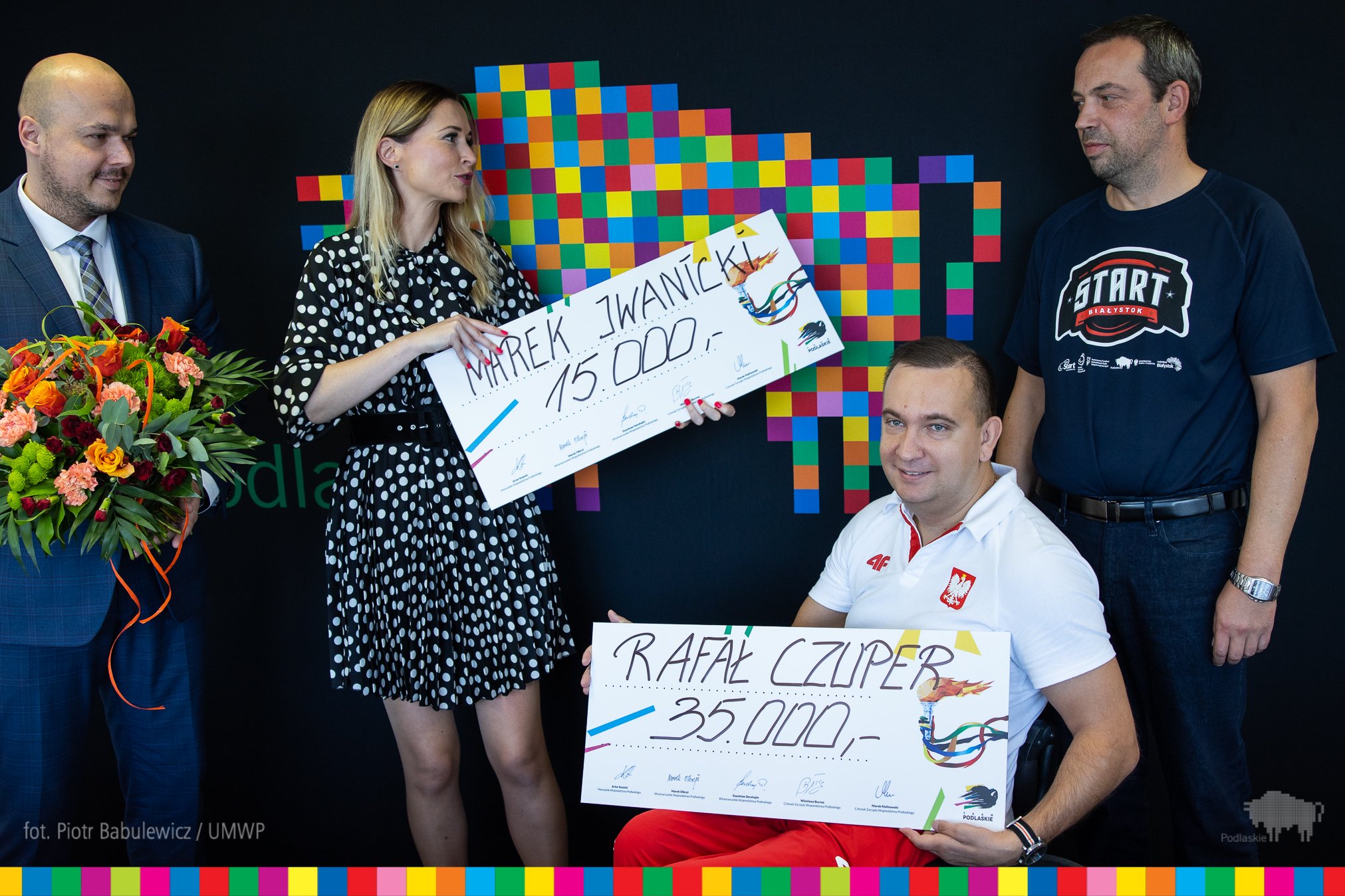 Nagrodzony sportowiec odbiera symboliczny czek, w tle stoi jego trener i przedstawiciele urzedu marszałkowskiego