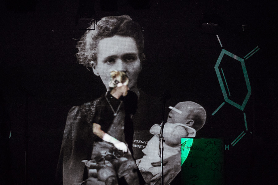 Kobieta na scenie, w tle wizualizacja przedstawiająca Marię Skłodowską-Curie