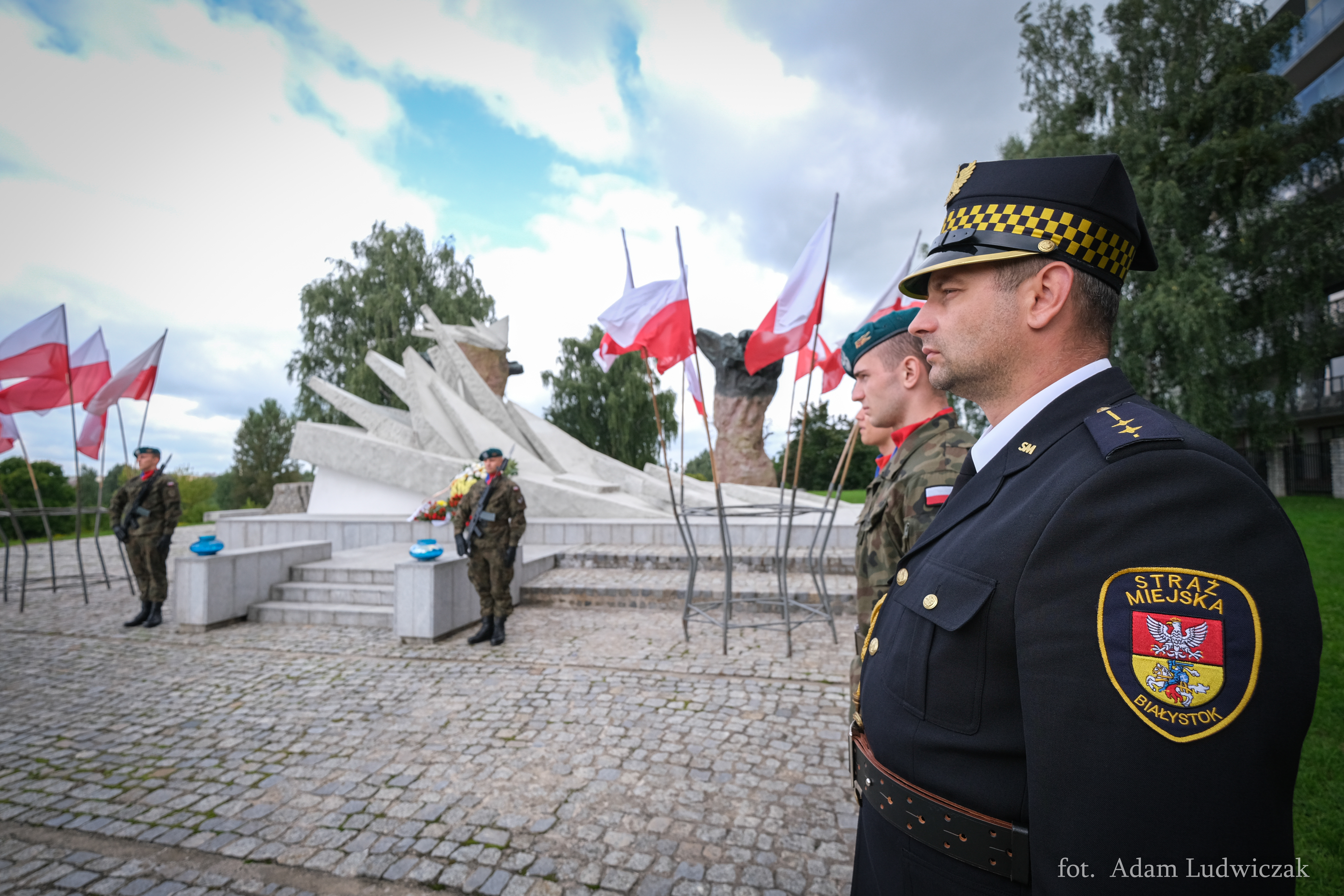 żołnierze i strażnicy miejscy stoją przy pomniku obrońców miasta, przed pomnikiem stoją znicze, na wietrze trzepoczą biało-czerwone flagi