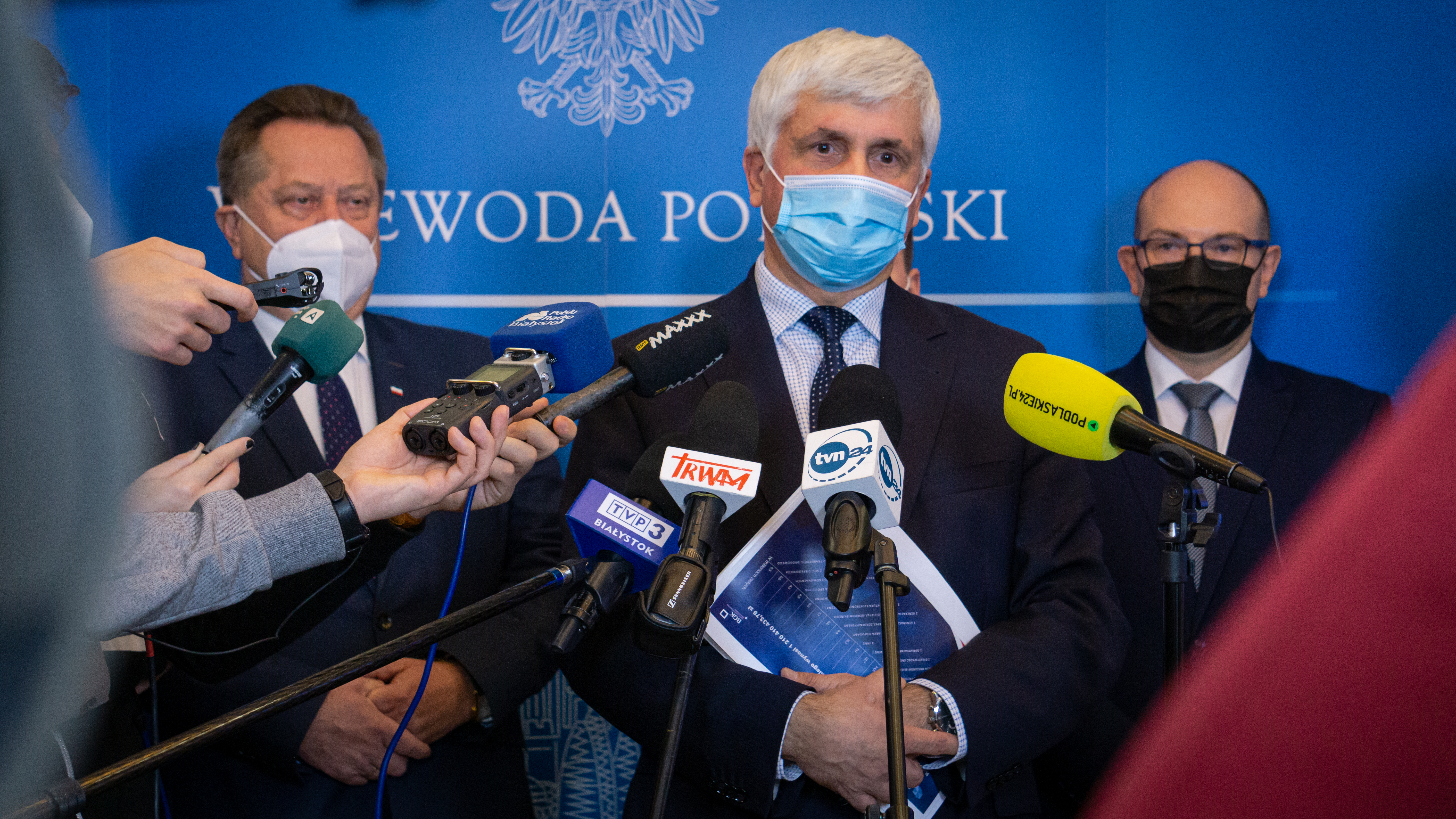Mężczyzna w ciemnym garniturze, w maseczce ochronnej na twarzy, o siwych włosach i brązowych oczach przemawia do dziennikarzy, do mikrofonów różnych redakcji, które stoją przed nim. Za mężczyzną na niebieskiej ściance jest biały napis wojewoda podlaski. O ściankę opiera się dwóch mężczyzn