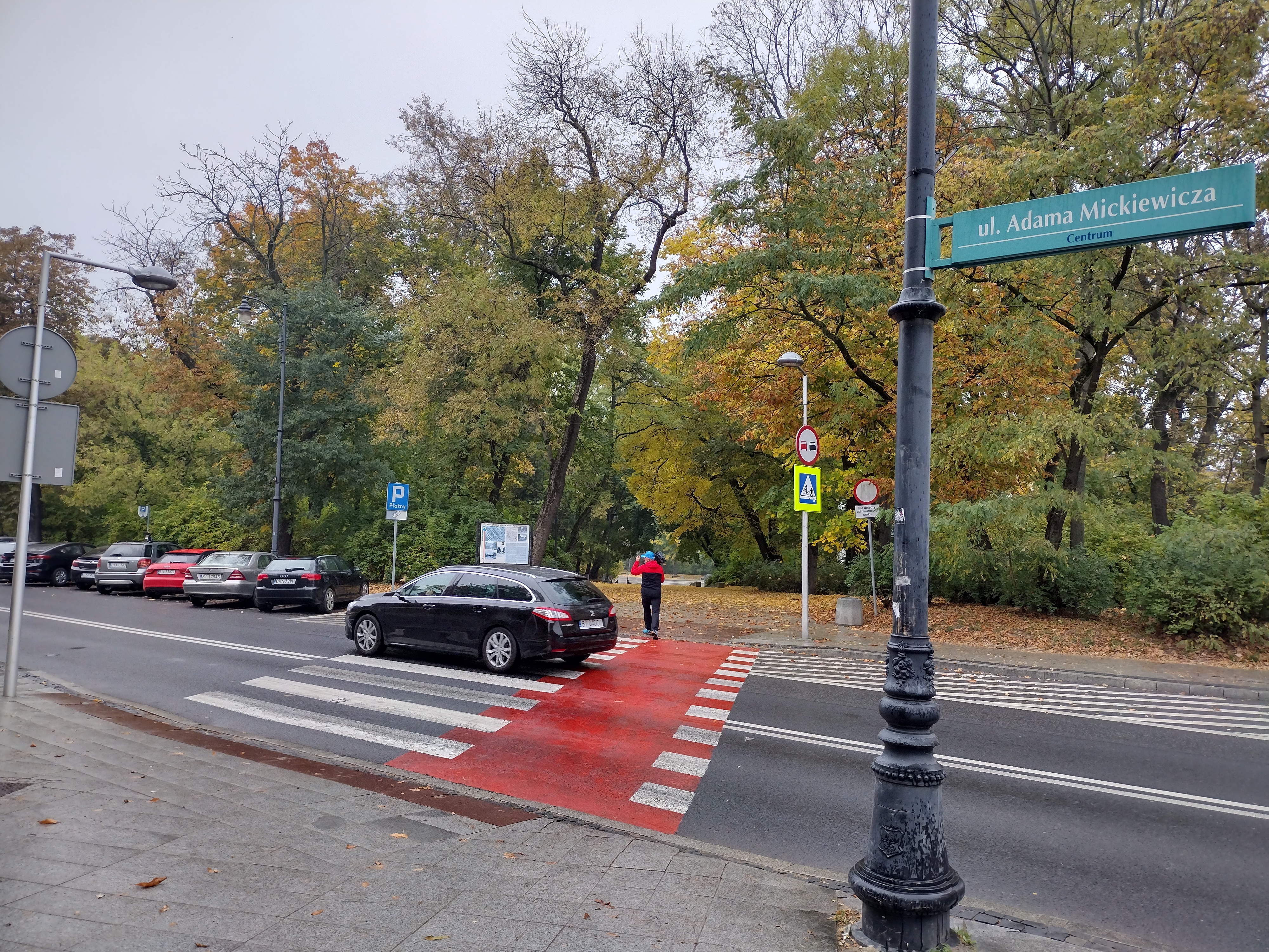 Jezdnia, na której jest przejście dla pieszych. Przez przejście przechodzi pieszy i przejeżdża czarny samochód. Na pierwszym planie, z prawej strony jest latarnia, do której przyczepiona jest zielona tabliczka z nazwą ulicy: Adama Mickiewicza