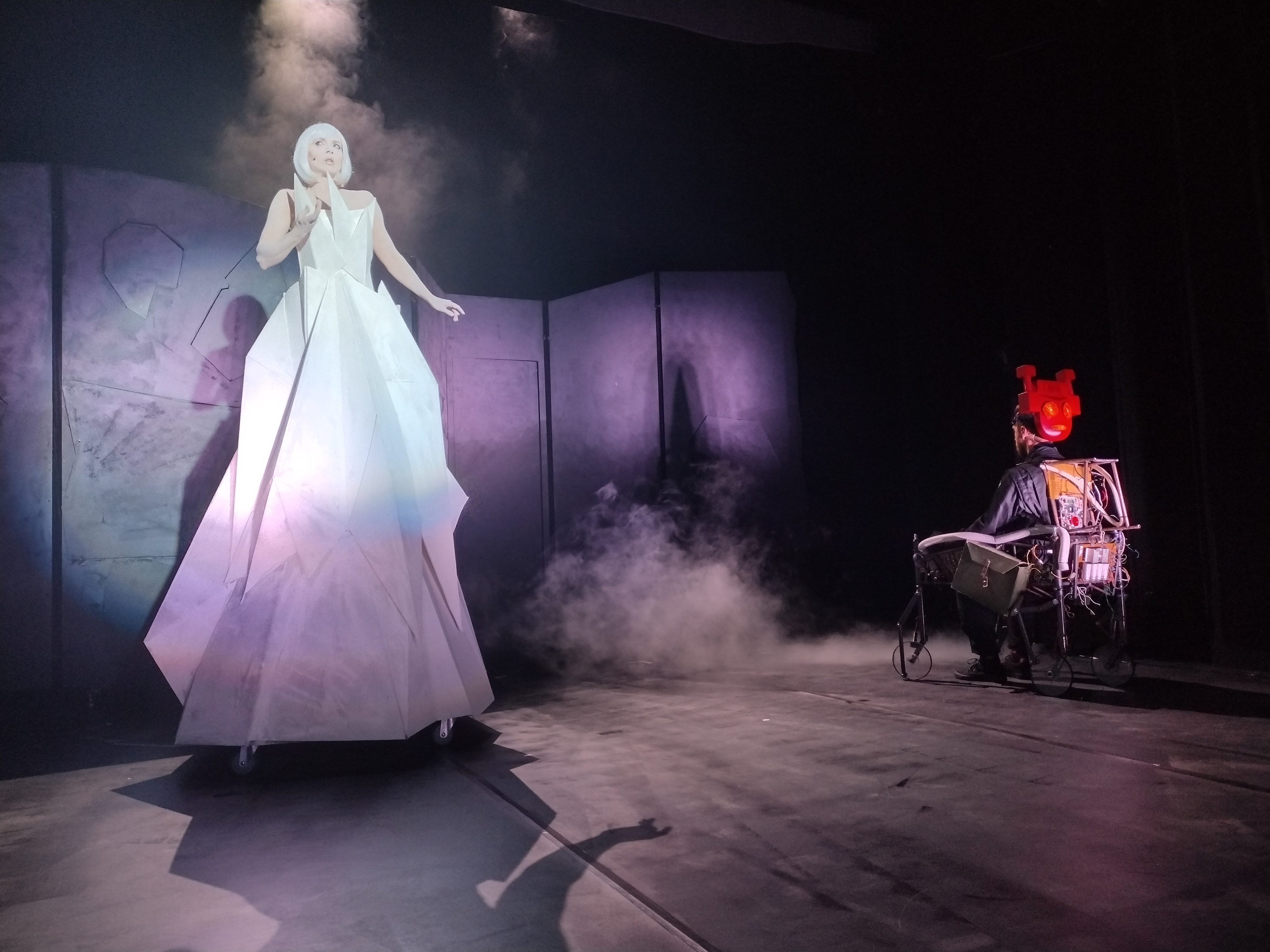 Scena teatralna, na której z lewej strony stoi Królowa Śniegu. Aktorka ma białe włosy i białą suknię. Kobieta jest bardzo wysoka, przemieszcza się na kółkach . Z prawej strony znajduje się robot, z czerwoną twarzą , która przemieszcza się na wózku