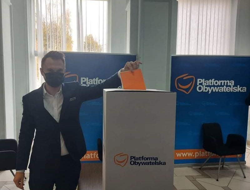 Mężczyzna w ciemnym garniturze i białej koszuli wrzuca kartkę do urny, która stoi obok niego. Urna jest biała z napisem: Platforma Obywatelska . Za mężczyzną stoją niebieskie banery z białym napisem: Platforma Obywatelska