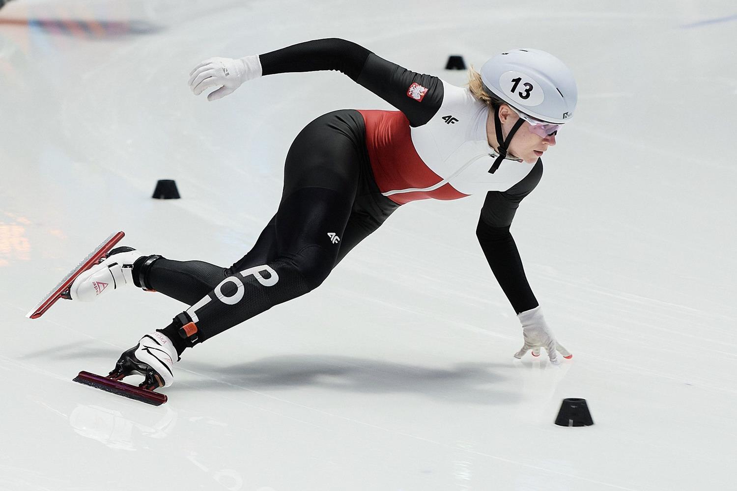 Łyżwiarka sportowa, specjalizująca się w short tracku jedzie po lodzie. Zawodniczka jest przechylona na lewą stronę, palcami dotyka do tafli. Kobieta jest ubrana w kombinezon, który do piersi jest biały, później czerwony, a od pasa czarny, rękawy też są czarne, na głowie ma biały kask