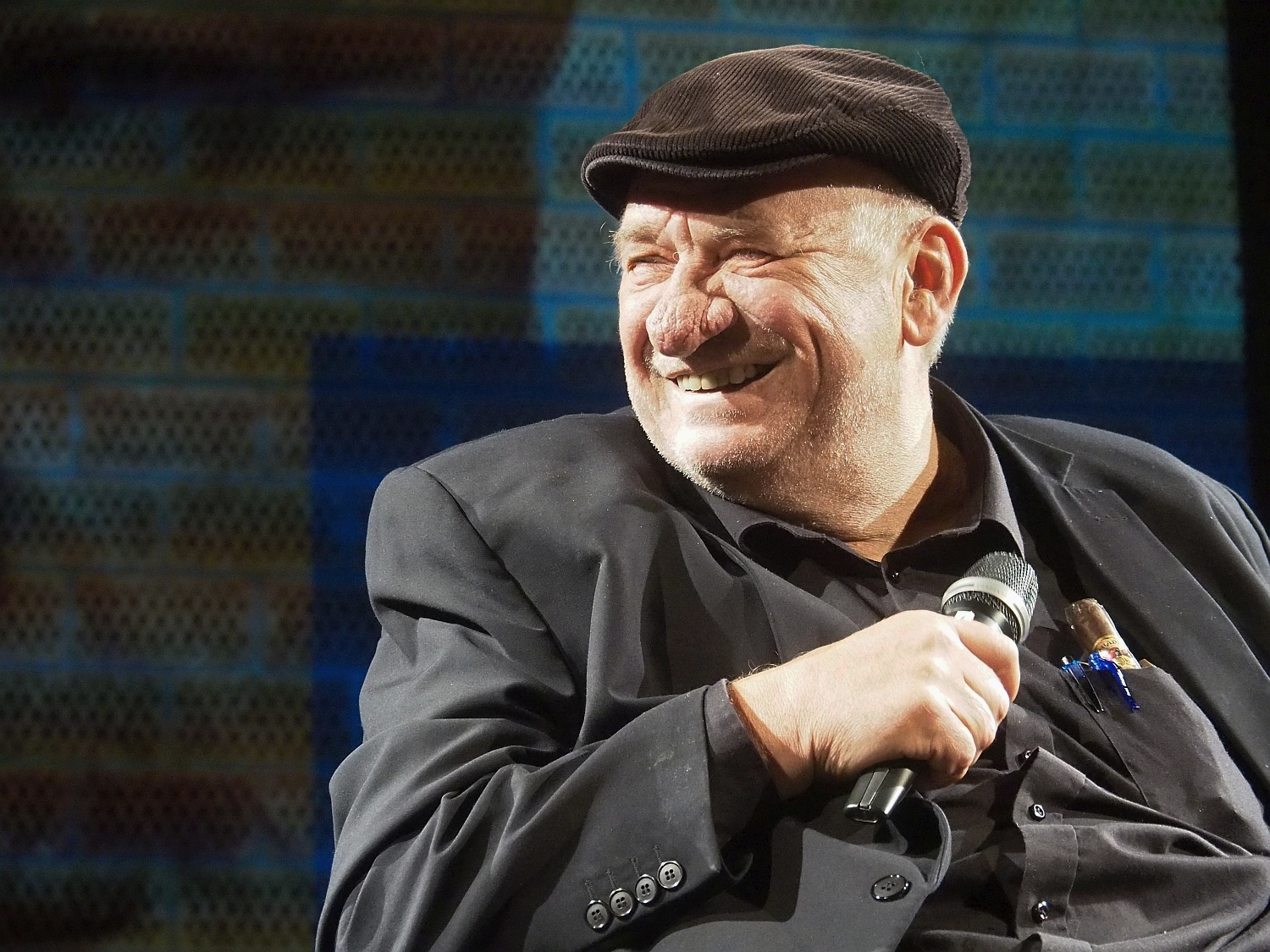 Ubrany na czarno mężczyzna siedzi na krześle, w dłoni trzyma mikrofon
