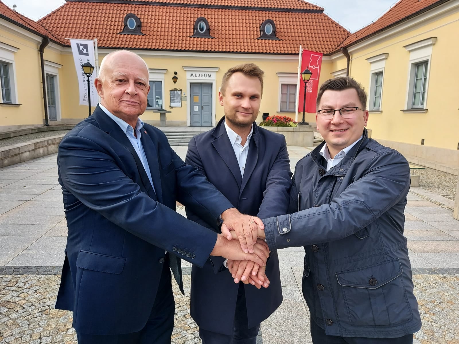 Trzech mężczyzn stoi obok siebie wykonując gest solidarności łącząc przed sobą dłonie. Mężczyźni stoją na tle białostockiego ratusza