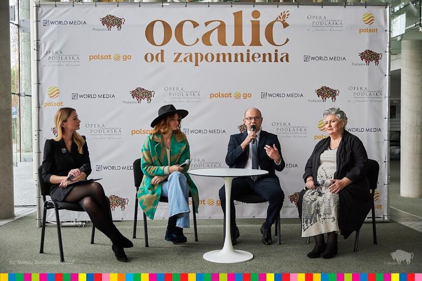 Na tle ścianki, na której jest złoty napis: Ocalić od zapomnienia siedzą cztery osoby, od lewej kobieta, kobieta, mężczyzna i kobieta. Siedzą na krzesłach przed nimi stoi biały, okrągły stoli. Mężczyzna mówi do mikrofonu, kobiety patrzą na niego
