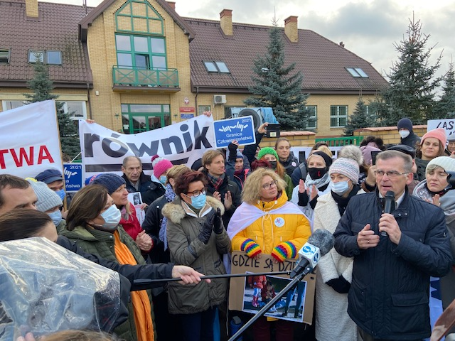 Grupa protestujących ludzi. W tłumie stoją pierwsze damy RP: Jolanta Kwaśniewska i Anna Komorowska, mężczyzna przemawia przez mikrofon. Za tłumem ludzi widoczny jest budynek z żółtej, klinkierowej cegły