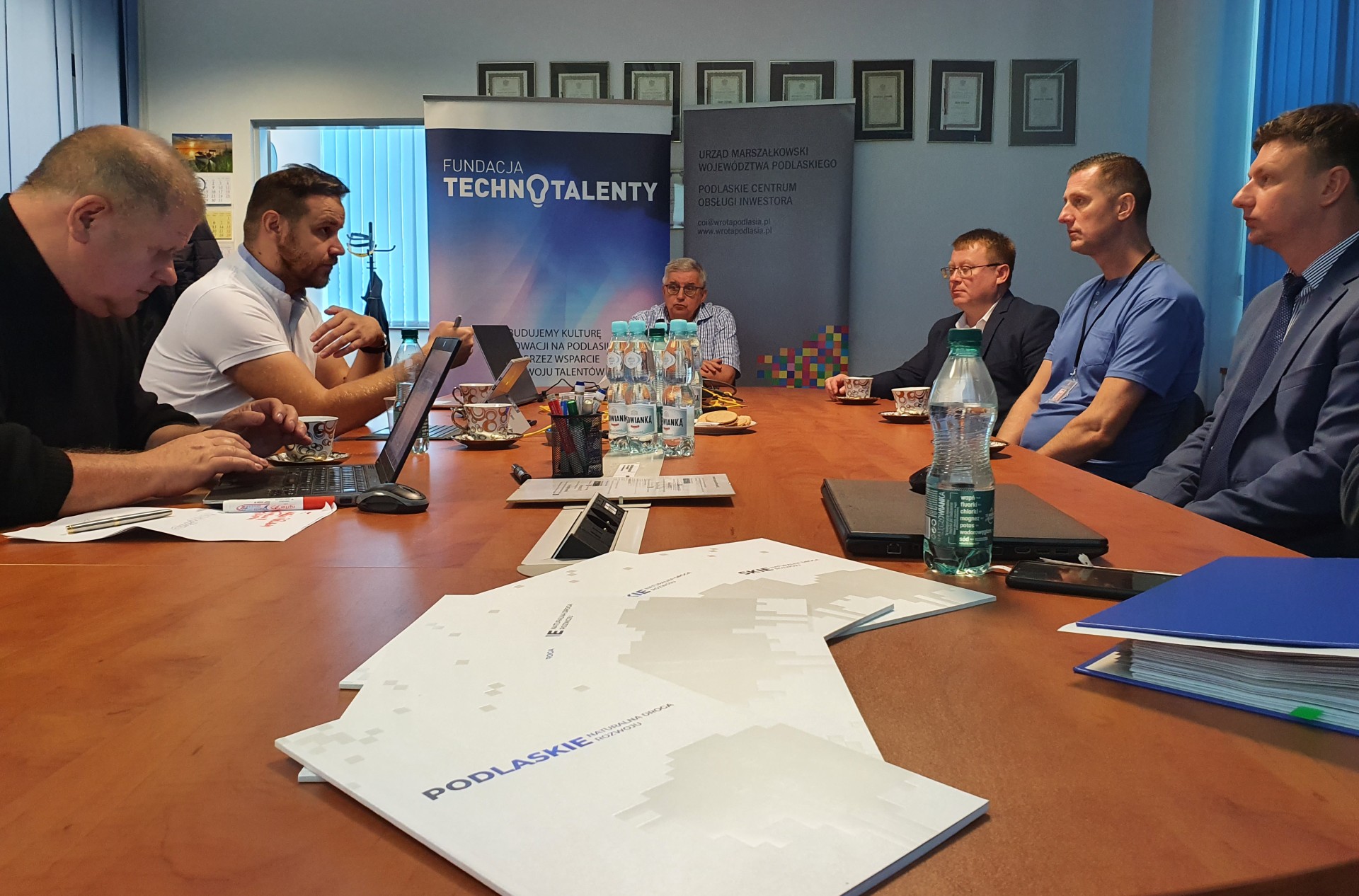 Mężczyźni siedzą przy brązowym stole, za nimi stoi baner z zapisem Fundacja Technotalenty