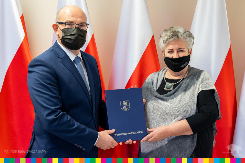 Mężczyzna w ciemny garniturze, w okularach i maseczce na twarzy przekazuje nominację dyrektorską kobiecie w czarno -szarym ubraniu , która ma maseczkę na twarzy