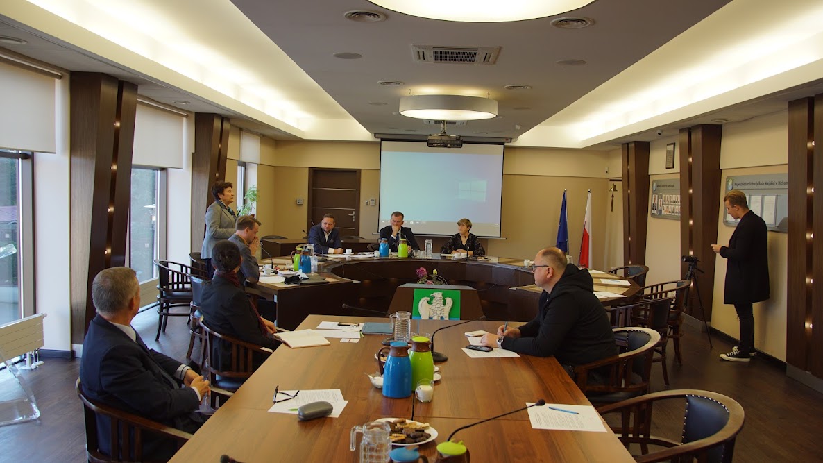 Sala konferencyjna, na środku której stoi prostokątny stół , przy którym siedzą ludzie. Na końcu sali pod ścianą stoi jeszcze jeden stół, w poprzek sali, przy nim również siedzą osoby. Za nimi znajduje się rzutnik