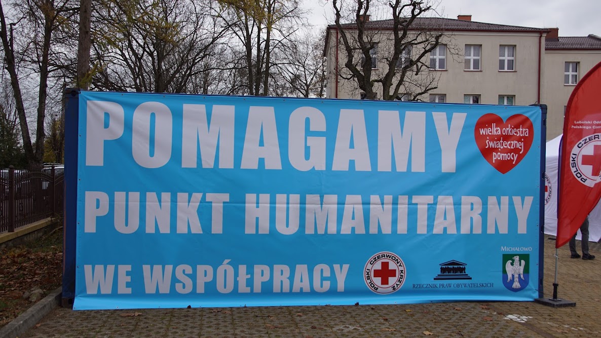 Błękitny, prostokątny baner z białym napisem: Pomagamy punkt humanitarny we współpracy, tu znajdują się loga Polskiego Czerwonego Krzyża, Rzecznika Praw Obywatelskich, gminy Michałowo, a w prawym górnym rogu Wielkiej Orkiestry Świątecznej Pomocy