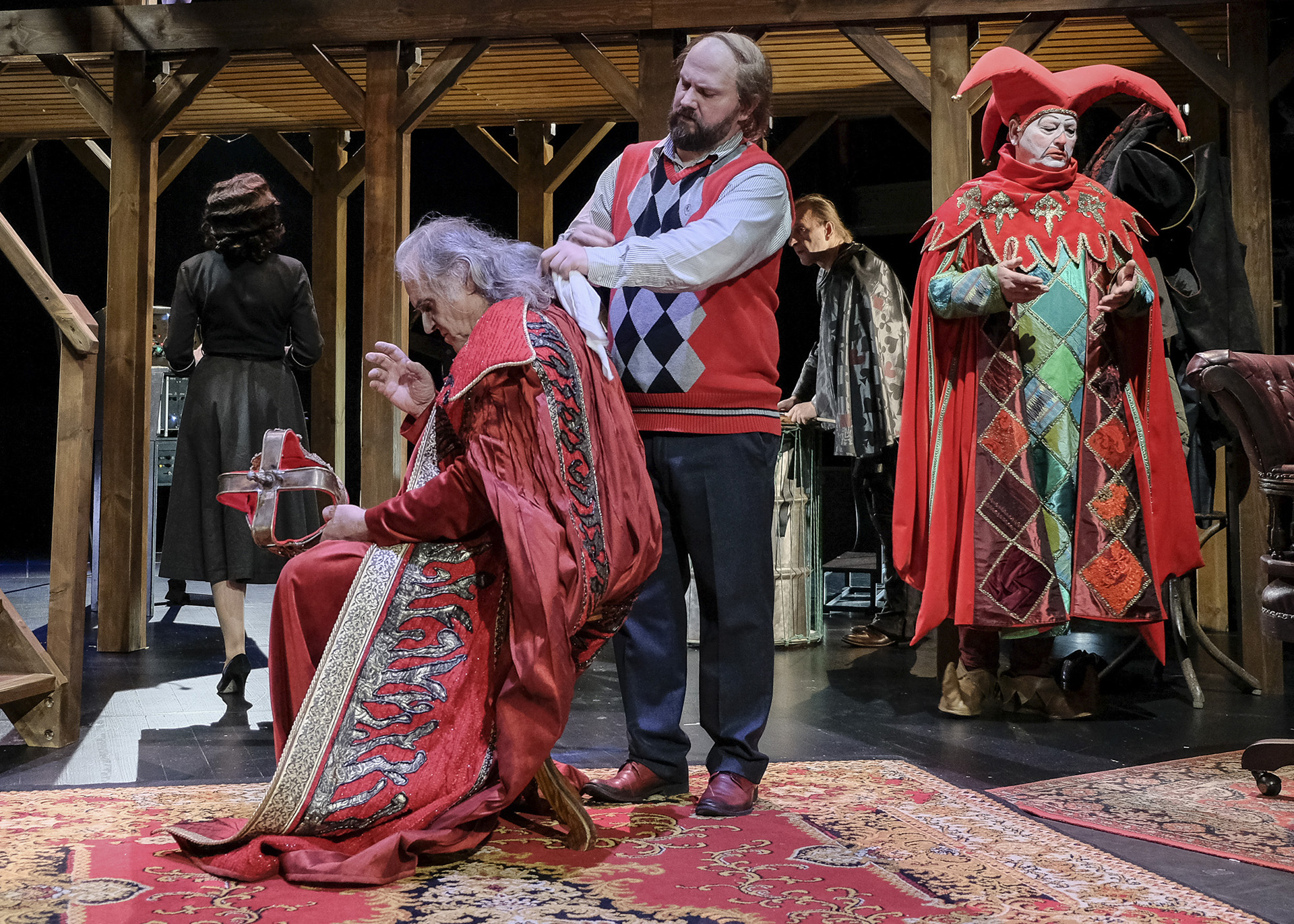 Scena teatralna, na podłodze leży czerwony, w roślinne wzory dywan, z lewej strony na krześle siedzi mężczyzna w czerwonym królewskim stroju, ma na sobie płaszcz heraldyczny, jego siwe włosy sięgające do szyi czesze mężczyzna, który stoi nad nim. To jest garderobiany, który ma na bok zaczesane włosy i jest ubrany w czerwony sweter w czarne romby na brzuchu. Z prawej strony stoi mężczyzna przebrany za błazna, ma kolorową pelerynę, w dominującym czerwonym kolorze, z przodu są zielone i bordowe romby. Ma na biało umalowaną twarz