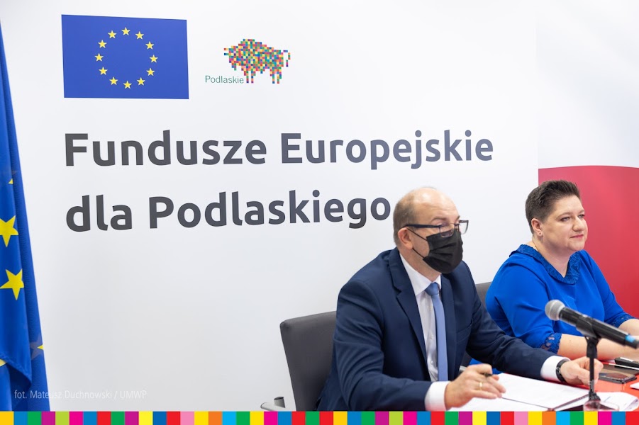 Mężczyzna i kobieta siedzą na tle białej ścianki, na której jest napisane czarnymi literami: Fundusze Europejskie dla Podlaskiego