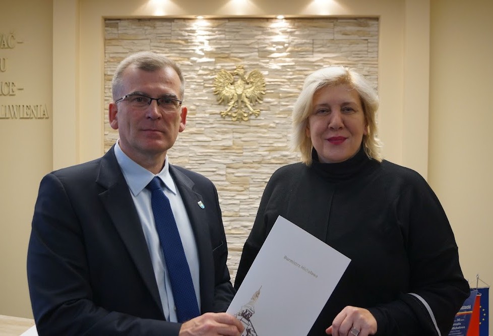 Na tle kremowej ściany, na której wisi złote godło Polski stoi mężczyzna w ciemnym garniturze i kobieta w czarnej bluzie. Mężczyzna trzyma przed sobą w ręku białą teczkę