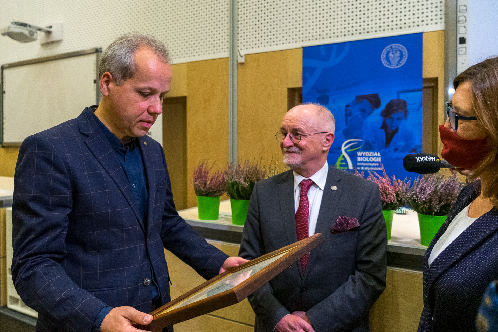 Mężczyzna, który stoi bokiem na zdjęciu, z lewej strony trzyma w rękach ramę, w którą oprawiony jest dyplom. Bliżej prawej strony stoi mężczyzna w szarym garniturze z bordowym krawatem