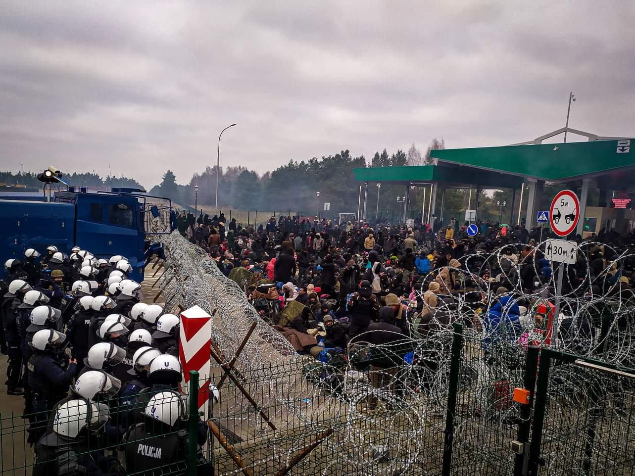 Kordon policji w białych kaskach stoi przed drutem kolczastym, który rozciągnięty jest na granicy