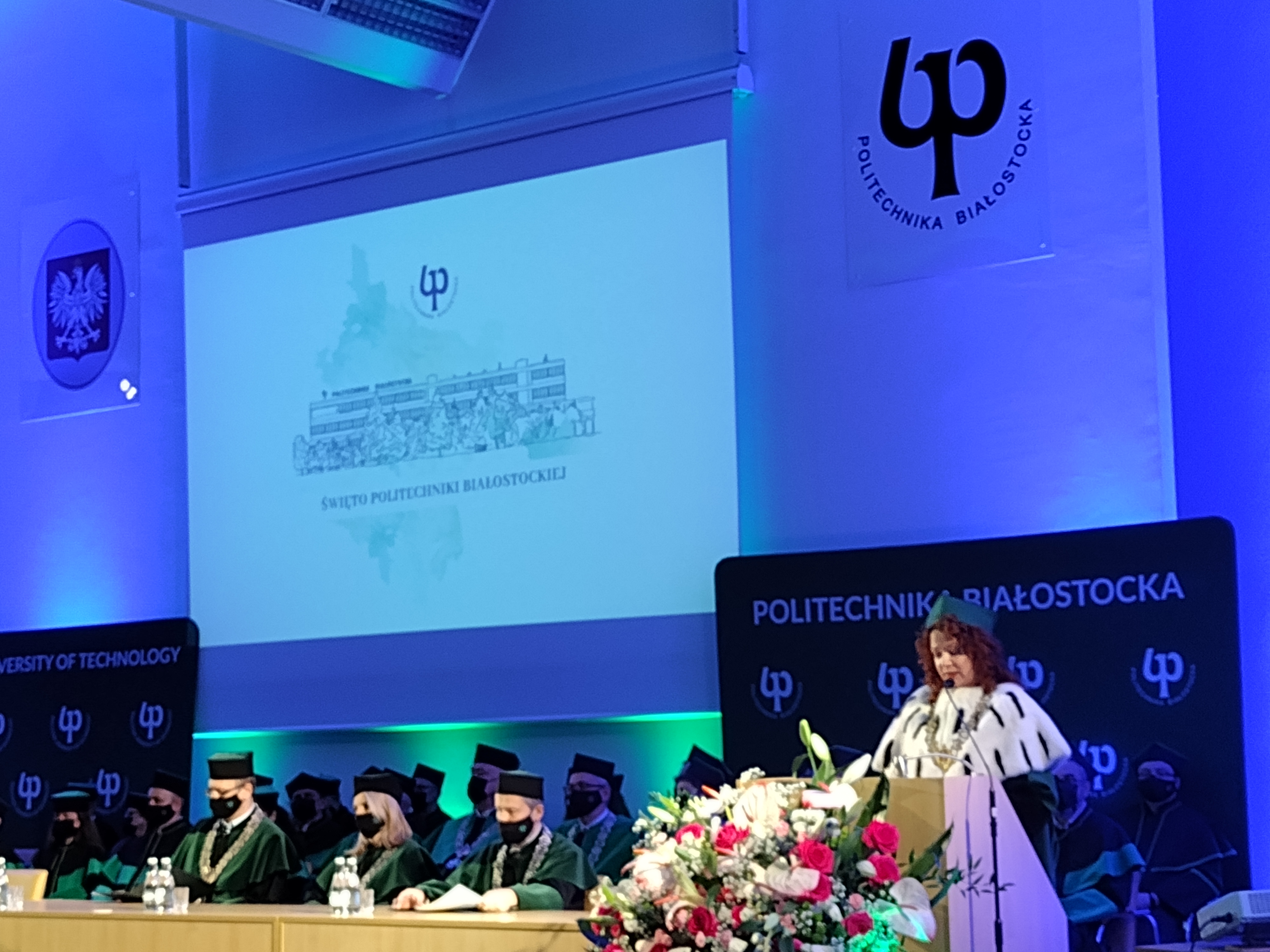 Fragment auli, przy mównicy stoi kobieta, za stołem siedzą osoby w czarno-zielonych togach. Nad nimi znajduje się rzutnik, na którym jest wyświetlony napis Święto Politechniki Białostockiej. Po prawej stronie na ścianie znajduje się znak Politechniki Białostockiej, z prawej godło Polski