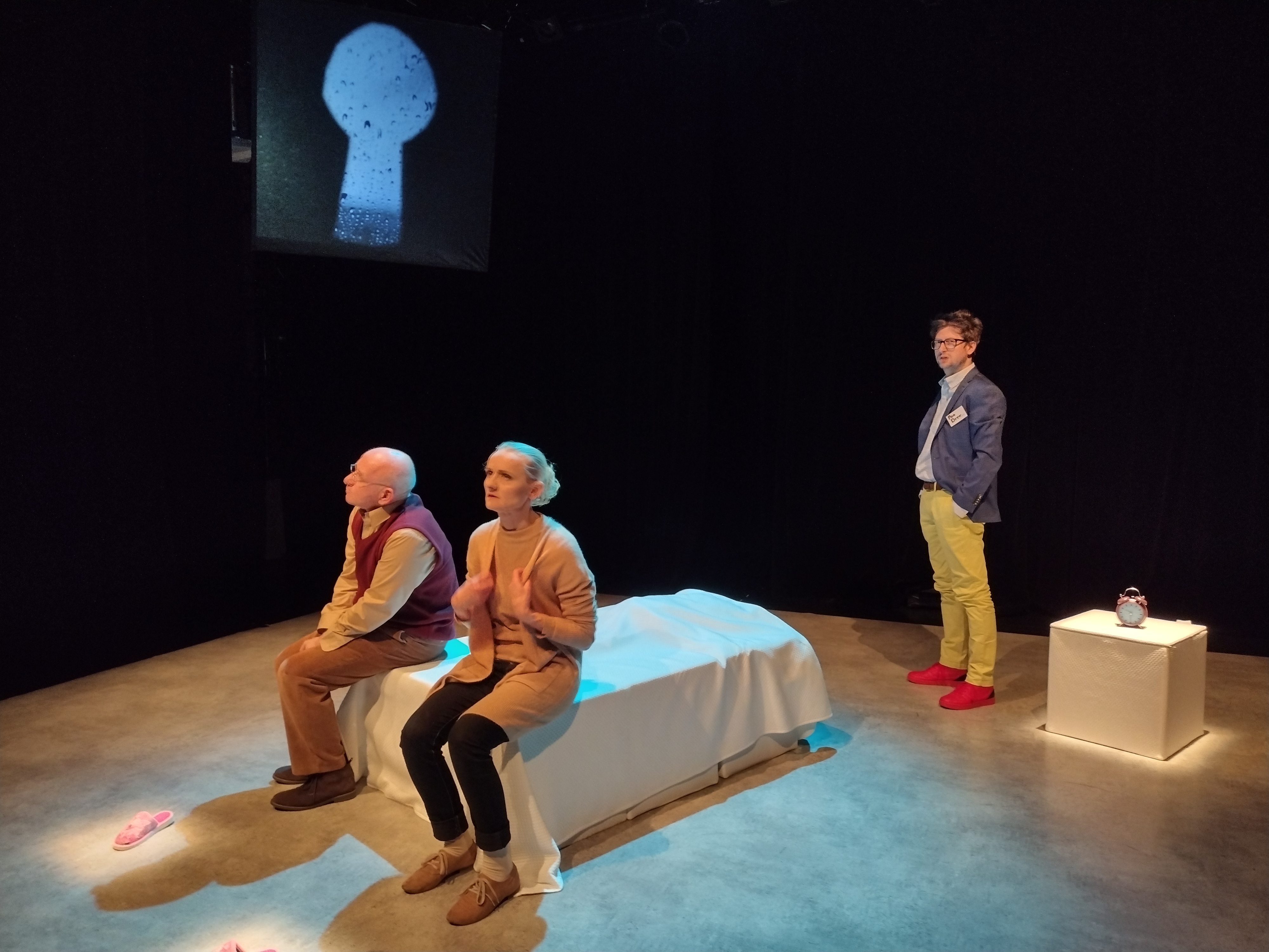 Scena teatralna, na łóżku siedzi kobieta i mężczyzna, na drugim planie planie stoi mężczyzna. Ciemne tło, z lewej strony na rzutniku wyświetlana jest dziurka od klucza przez, którą widać, że pada deszcz