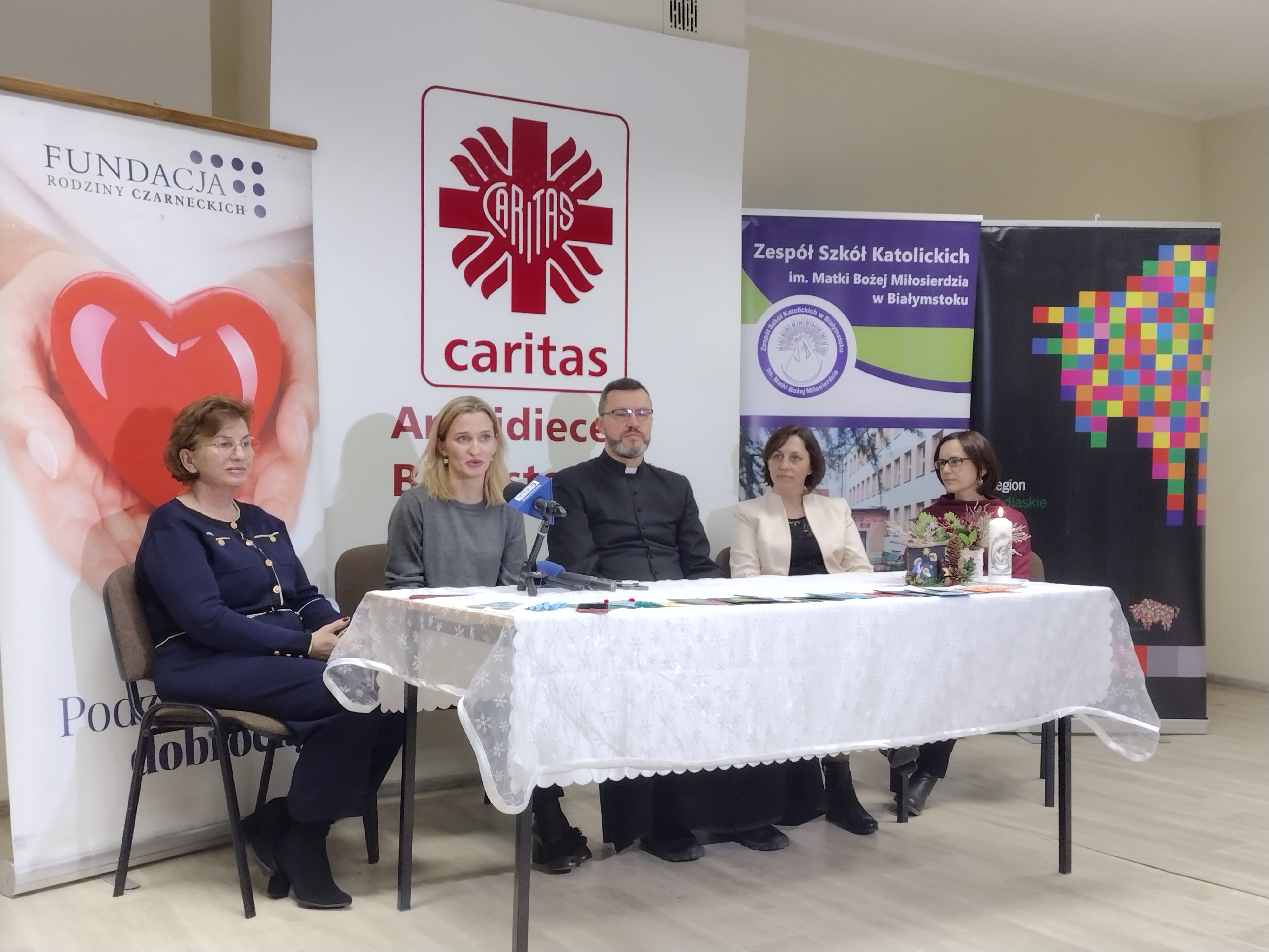 Przy stole nakrytym białym obrusem siedzi pięć osób. Po dwie kobiety po bokach, w środku ksiądz. Za nimi stoją banery; Fundacji Rodziny Czarneckich, Caritas Polska, województwa podlaskiego