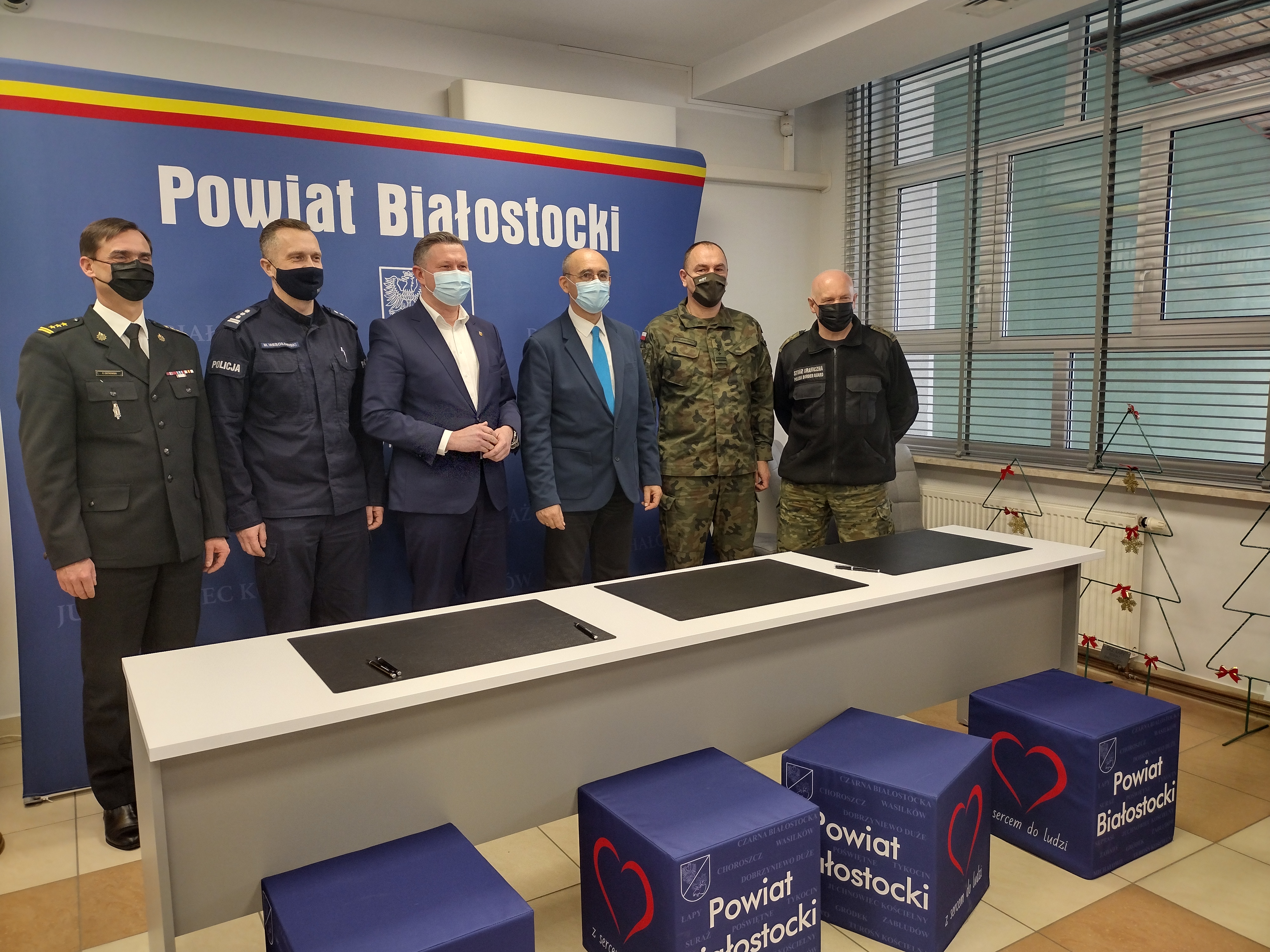 Na niebieskiej ściance z białym napisem Powiat białostocki. Na jej tle stoi sześciu mężczyzn; od lewej przedstawiciel strazy pożarnej, następnie policji, starosta białostocki, wicestarosta białostocki, przedstawiciel Wojsk Obrony Terytorialnej oraz Straży Granicznej