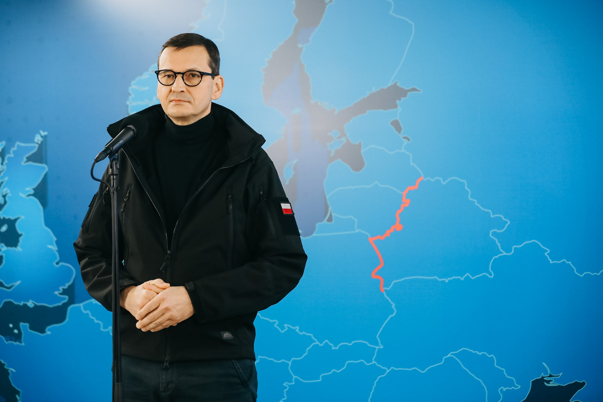 Na niebieskim tle stoi ubrany na czarno mężczyzna, który ma ciemne włosy i okulary lecznicze w ciemnych oprawkach. Mężczyzna stoi przed mikrofonem, do którego mówi.