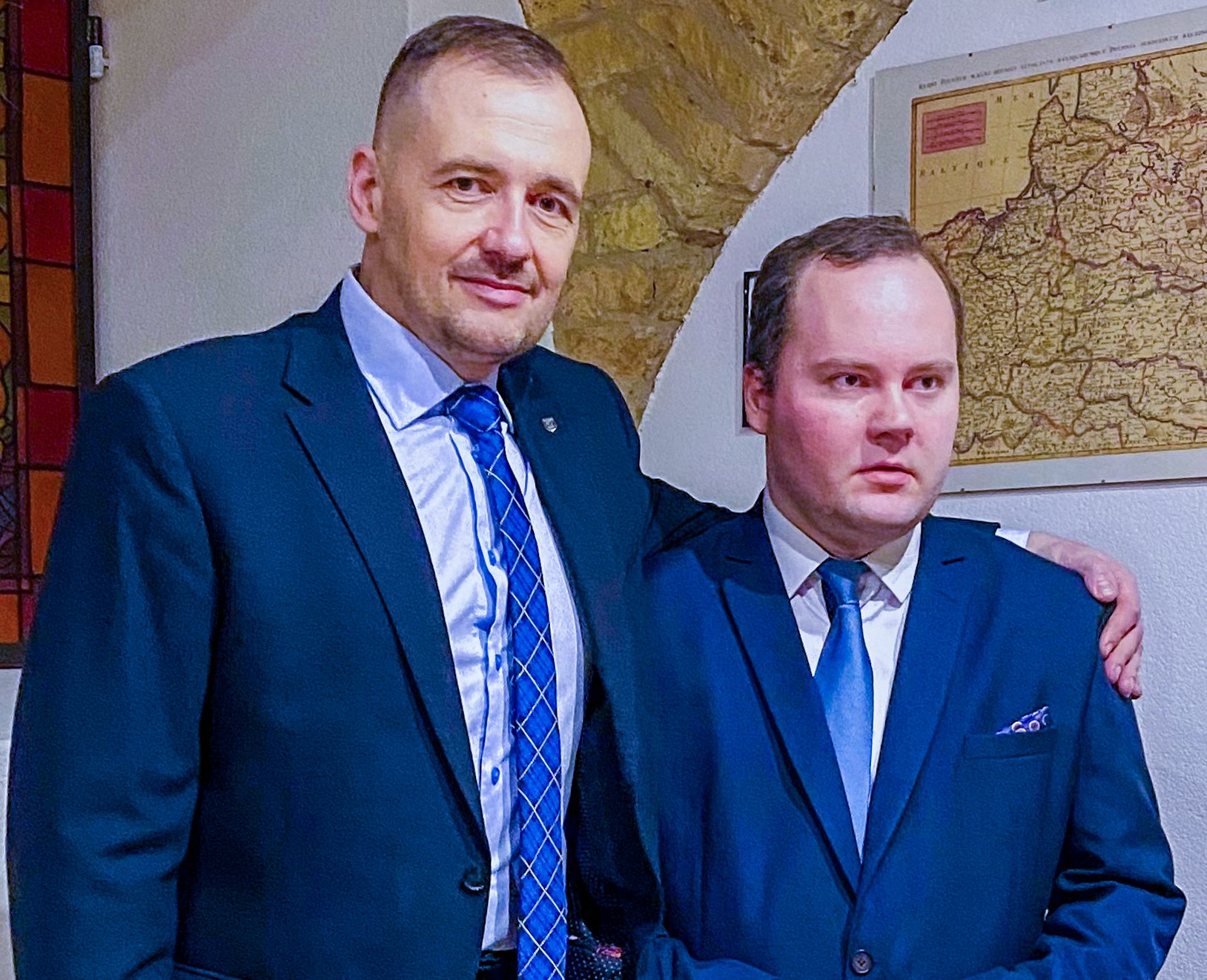 Dwóch mężczyzn w granatowych garniturach stoi obok siebie. Mężczyzna z lewej strony jest wyższy i obejmuje ręką stojącego obok siebie mężczyznę
