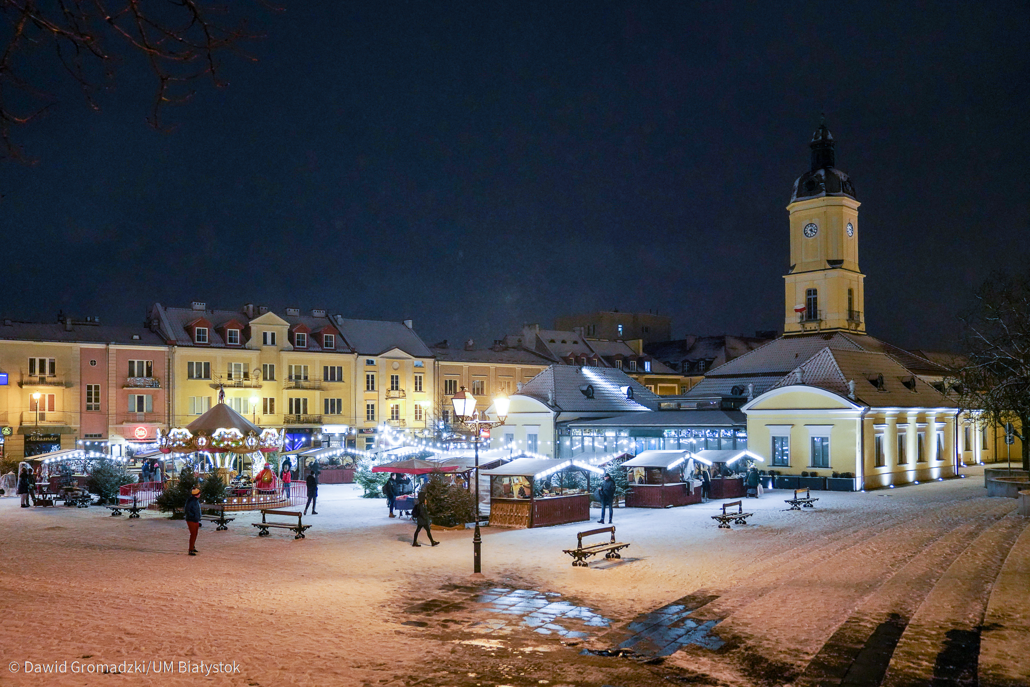 Noc, panoramiczne zdjęcia Rynku Kościuszki w Białymstoku. Z prawej strony stoi Ratusz, w oddali widać oświetlone drewniane, stragany Jarmarku Bożonarodzeniowego. Na trzecim planie widoczne są kamieniczki przy ul. Lipowej