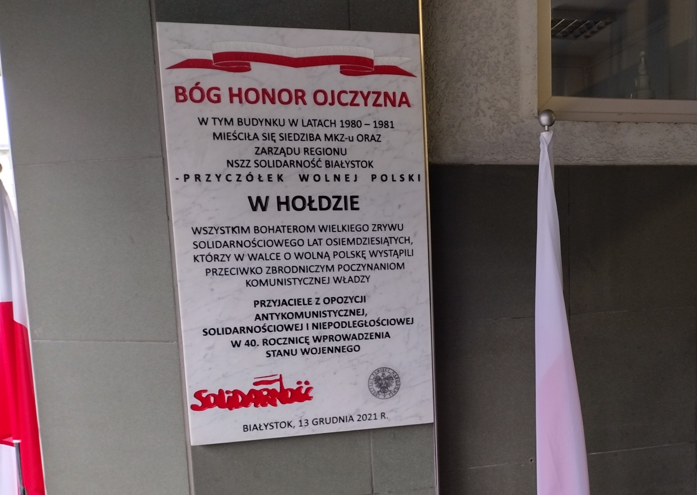 Biała, kamienna tablica z czarnymi napisami wisi na zewnętrznej ścianie. Po obu stronach tablicy znajdują się biało-czerwone flagi