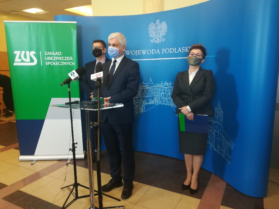 Trzy osoby, kobieta i dwóch mężczyzn stoi na tle banera Podlaskiego Urzędu Wojewódzkiego podczas konferencji prasowej. To: Bohdan Paszkowski – wojewoda podlaski, Piotr Jurczuk – zastępca dyrektora Izby Administracji Skarbowej w Białymstoku, Beata Pawlińska-Wiak – dyrektor oddziału Zakładu Ubezpieczeń Społecznych w Warszawie