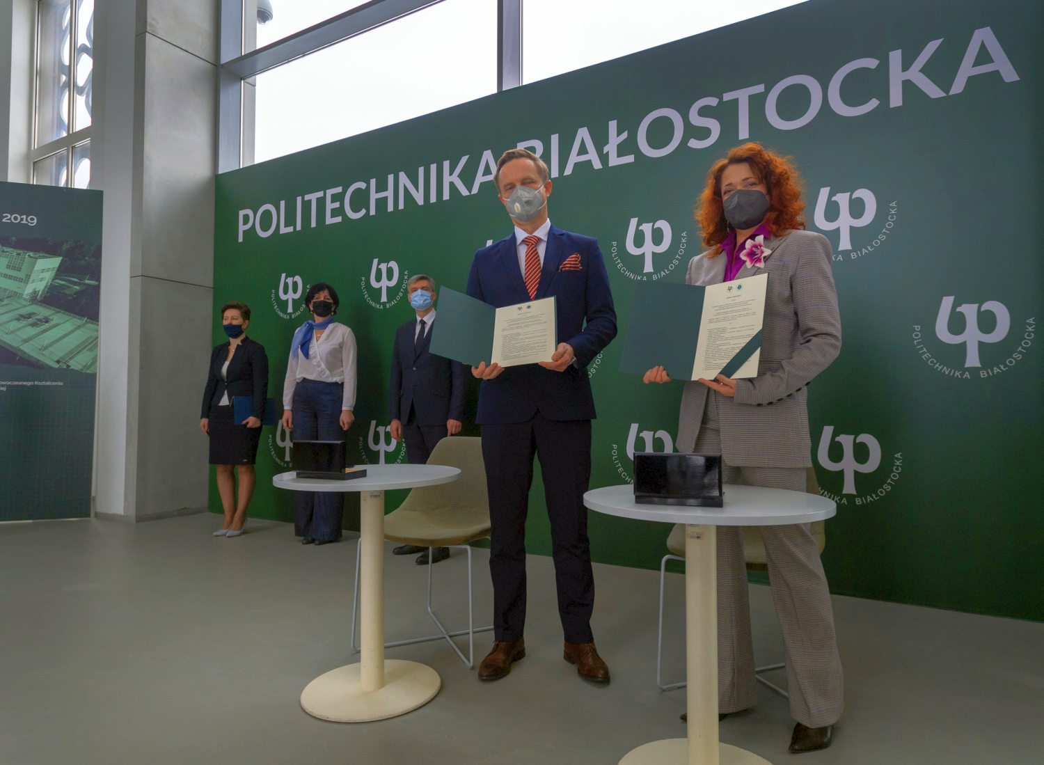 Na zielonej ściance z białym napisem: Politechnika Białostocka, z prawej strony stoi kobieta, z lewej mężczyzna. Oboje stoją przy dwóch białych okrągłych stolikach i trzymają przed sobą podpisane porozumienie o współpracy