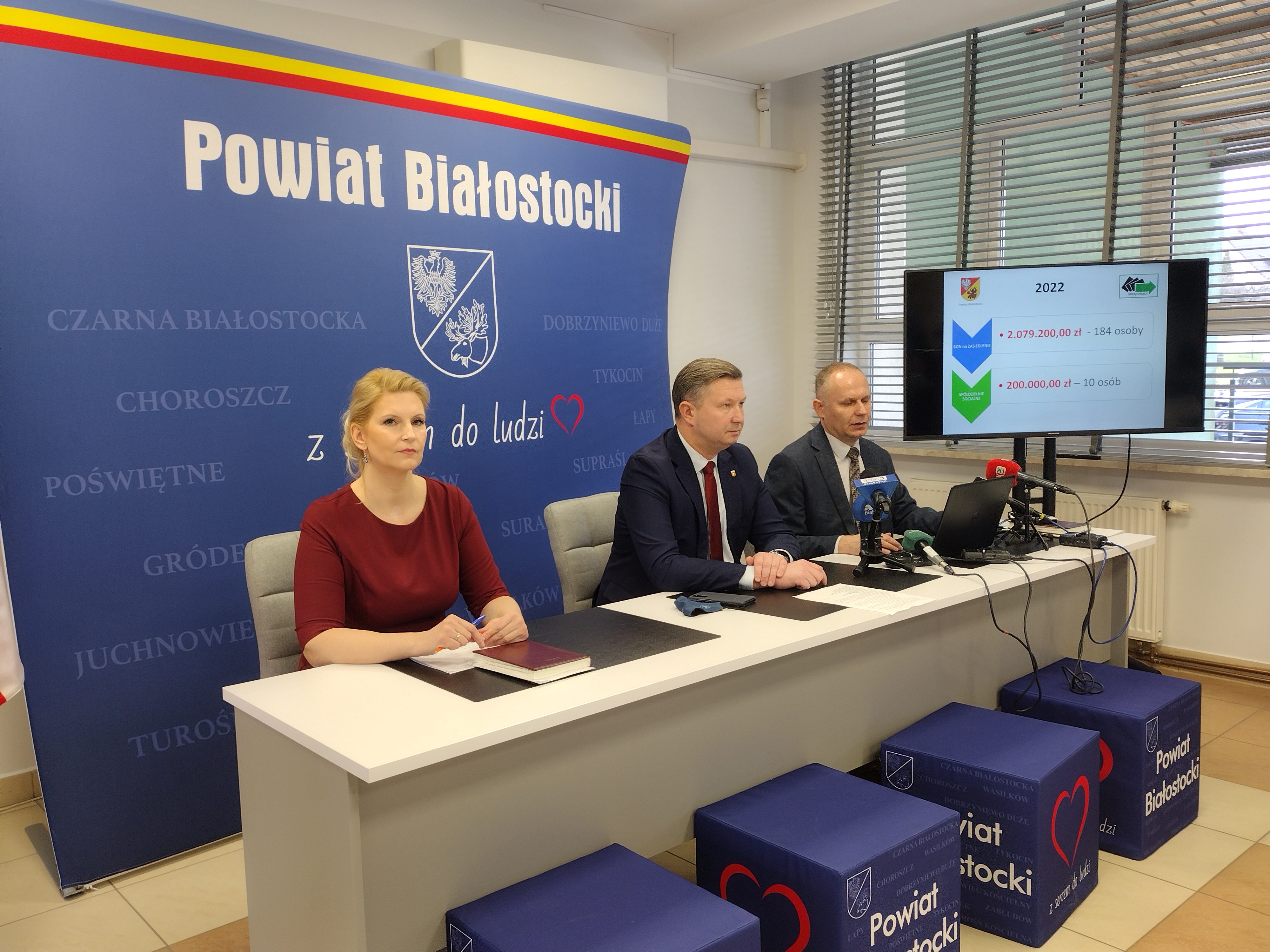 Przy jasnym stole siedzą trzy osoby na tle niebieskiej ścianki z białym napisem: Powiat Białostocki. Od lewej strony siedzi kobieta w bordowej sukience, po środku i z lewej strony mężczyźni w ciemnych garniturach. Z prawej strony stoi monitor, na którym wyświetlana prezentacja