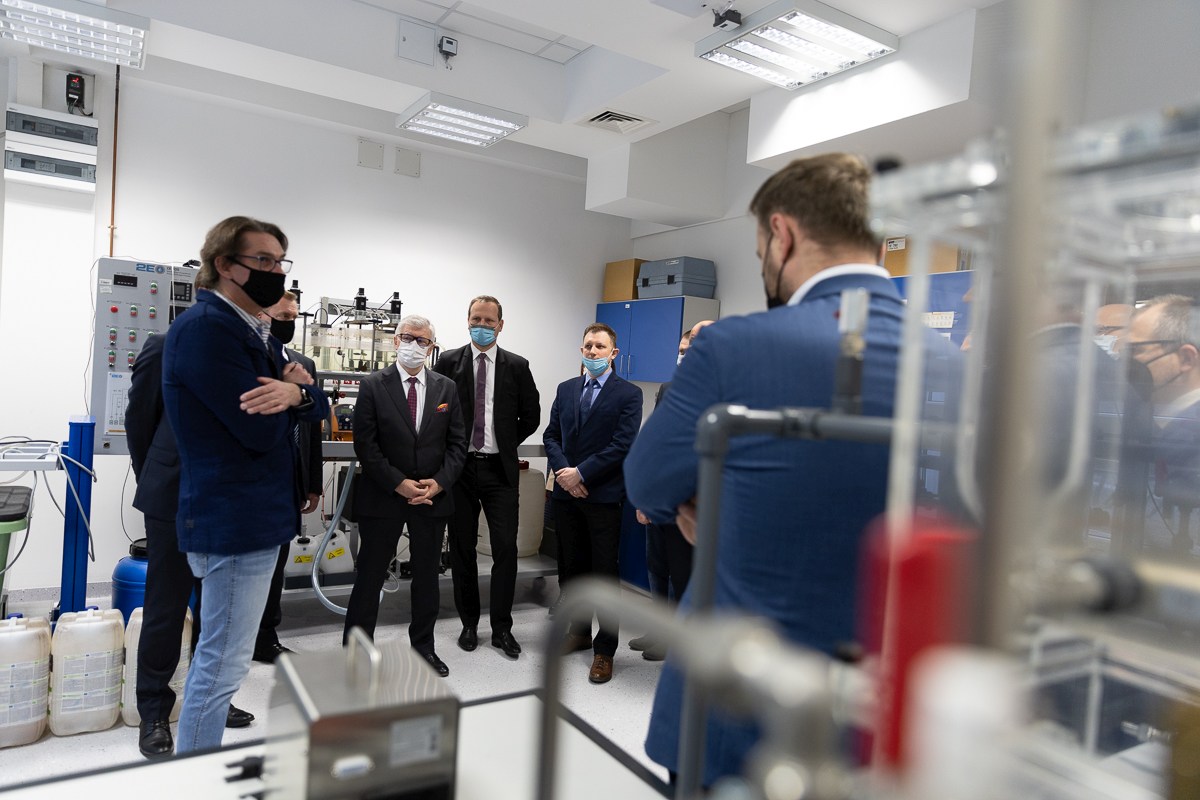 W laboratorium stoi grupa mężczyzn ubrana w ciemne garnitury. Mężczyźni słuchają, co mówi do nich inny mężczyzna