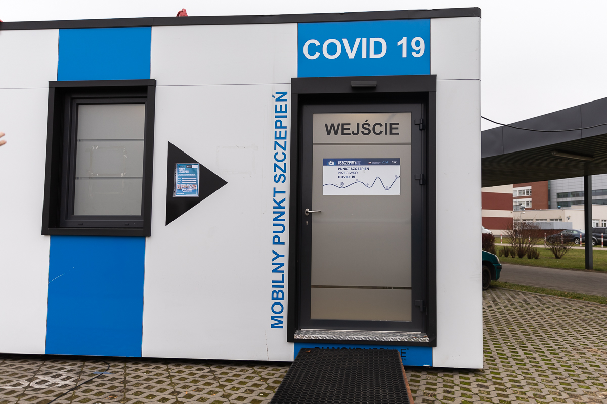 Wejście do konternerowego mobilnego punktu szczepień przeciwko COVID-19. Fragment punktu jest w biało-niebieskie pionowe pasy. Drzwi wejściowe są szare, nad nimi znajduje się biały napis na niebieskim tle: COVID-19