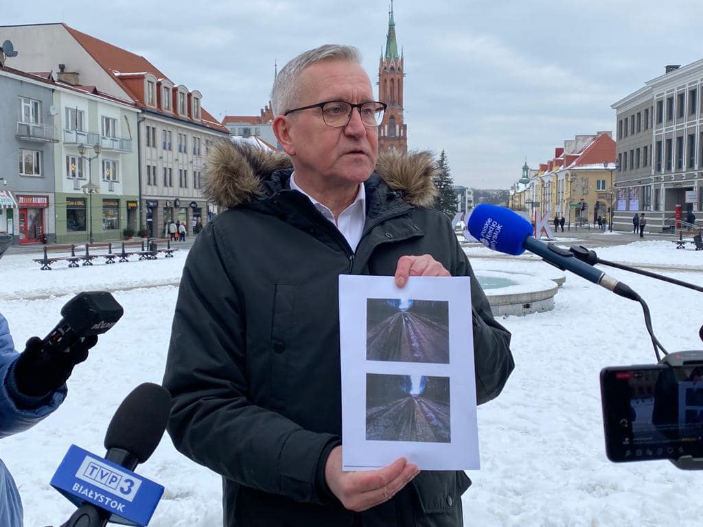 Mężczyzna w ciemnej kurtce z kapturem obszytym futrem stoi w centrum Białegostoku, za nim w oddali widać wieżę Katedry. Poseł mówi do mikrofonów zgromadzonych wokół niego dziennikarzy. Mężczyzna trzyma przed sobą zdjęcie zniszczonej drogi