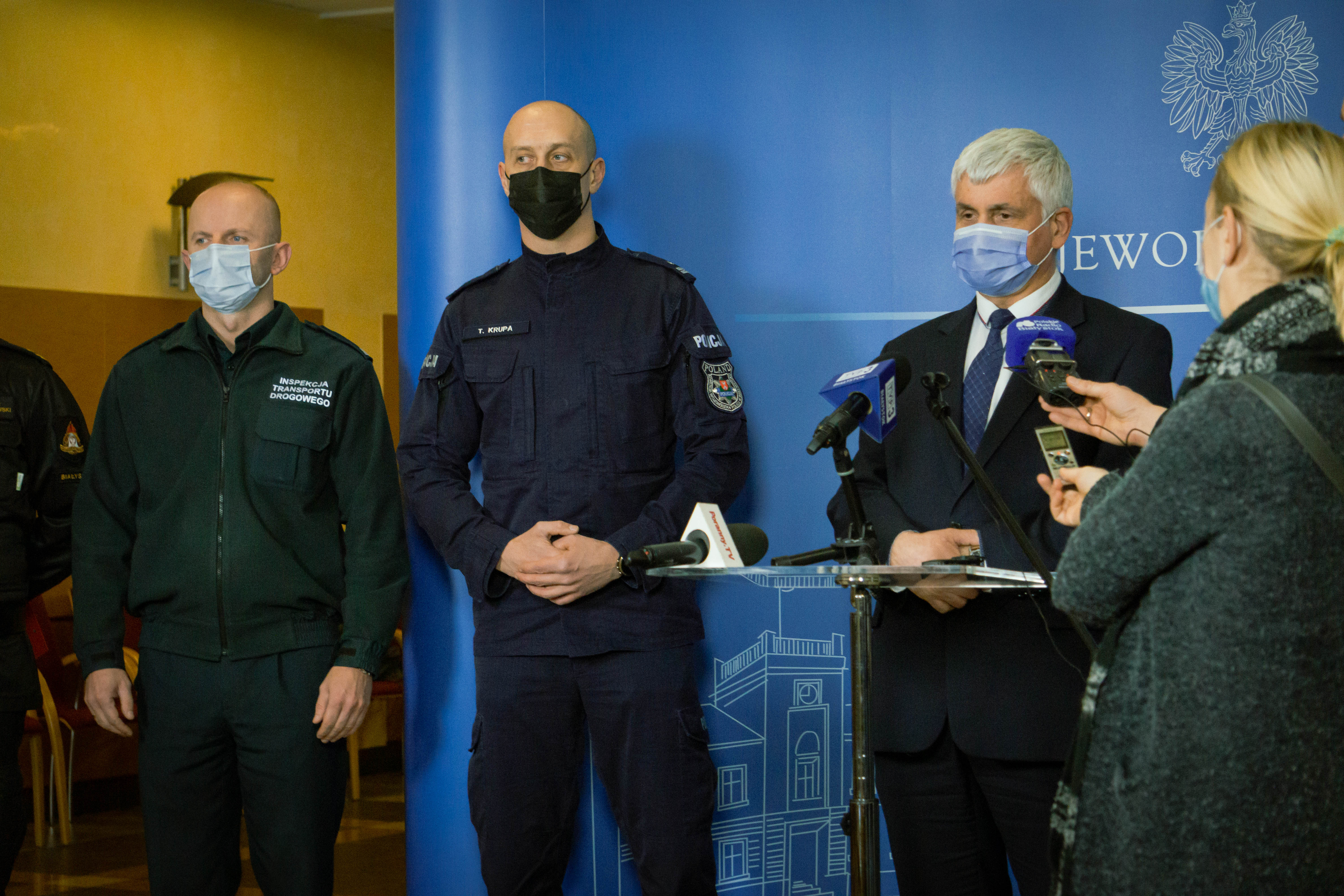 Na niebieskiej ściance stoją trzy osoby z prawej strony przy szklanym stoliku wojewoda, po środku stoi policjant w granatowym mundurze, z lewej przedstawiciel Inspekcji Transportu Drogowego
