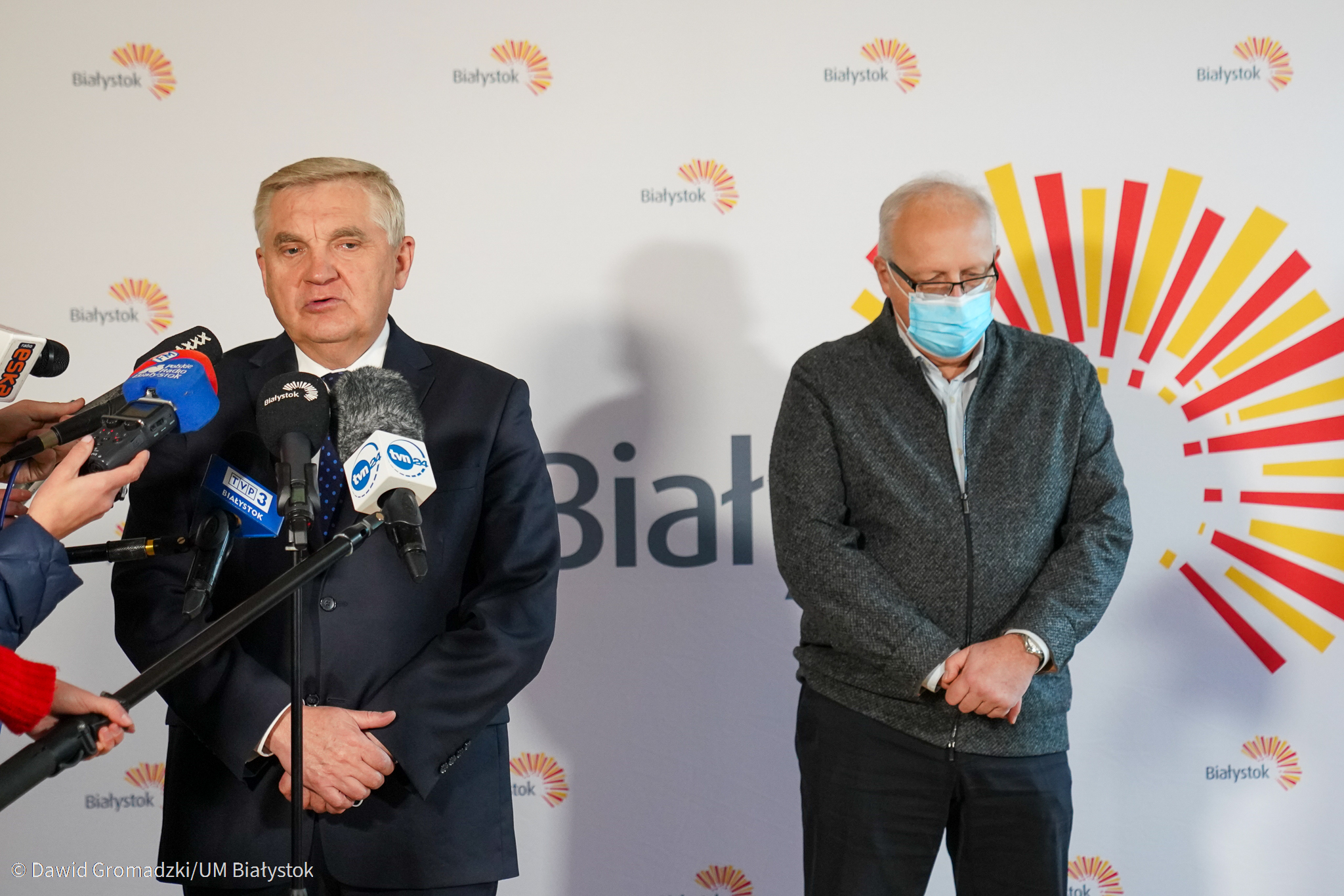 Na tle białej ścianki z napisem Białystok stoi dwóch mężczyzn. Z lewej prezydent Białegostoku, w ciemnym garniturze mówi do mikrofonów. Z prawej stoi mężczyzna ze spuszczoną głową, w szarym swetrze i w ciemnych spodniach