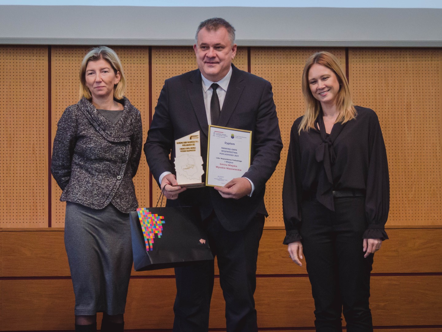 Na podeście w sali konferencyjnej stoją trzy osoby. Po środku mężczyzna, który trzyma przed sobą dyplom i statuetkę., po jego obu stronach znajdują się kobiety