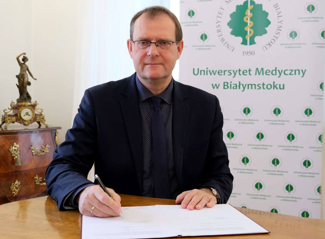 Mężczyzna w ciemnej koszuli i marynarce siedzie przy stole i podpisuje położone przed sobą dokumenty, patrzy w obiektyw. Za nim, z prawej strony stoi rolla-up z napisem: Uniwersytet Medyczny w Białymstoku. Z lewej strony, za mężczyzną znajduje się brązowa, antyczna komoda, na której stoi złoty zegar z figurką