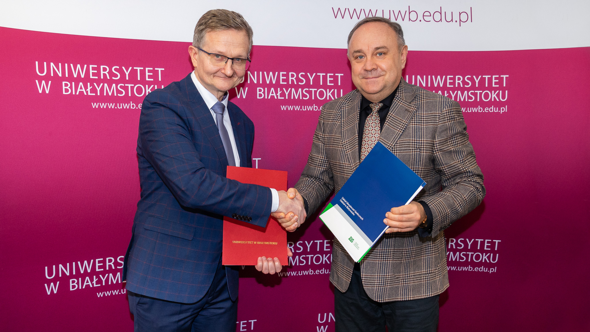 Na różowej ściance z białym napisem: Uniwersytet w Białymstoku stoi dwóch mężczyzn, którzy wzajemnie ściskają sobie dłonie