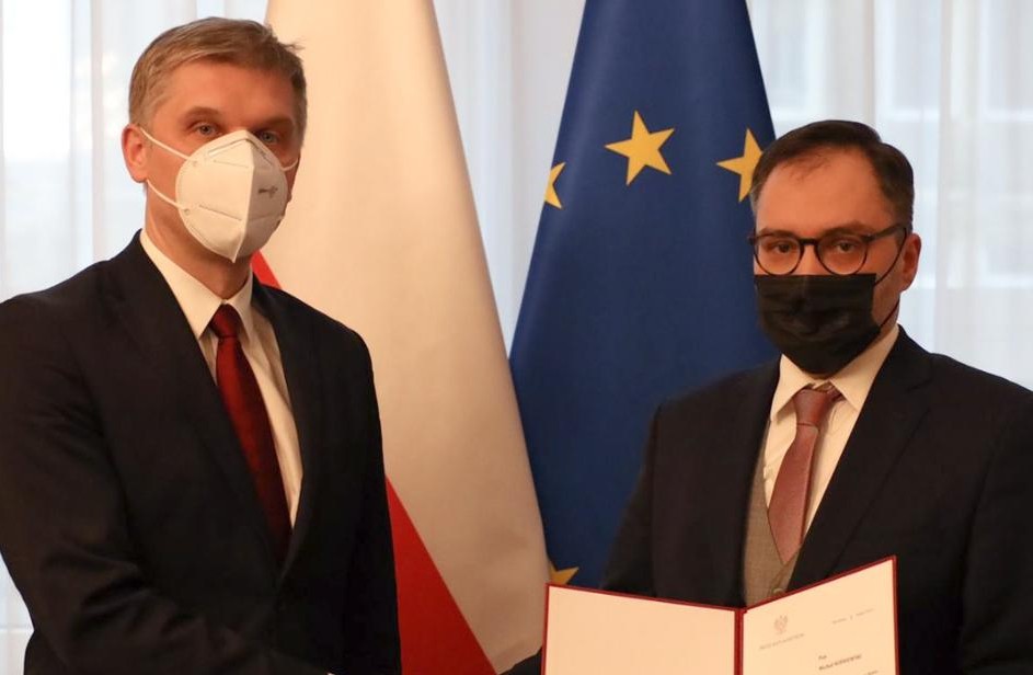 Dwóch mężczyzn w maseczkach ochronnych na twarzy stoi na tle Polski, biało-czerwonej oraz Unii Europejskiej, granatowej z żółtymi gwiazdkami w kręgu. Mężczyzna z prawej strony trzyma akt powołania