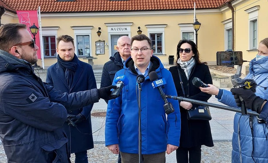 W centralnej części zdjęcia stoi mężczyzna w niebieskiej kurtce, mówi on do mikrofonów dziennikarzy stojących wokół niego. Za nim stoją trzy osoby, od lewej strony dwóch mężczyzn i kobieta. Na ostatnim planie widać fragment białostockiego Ratusza