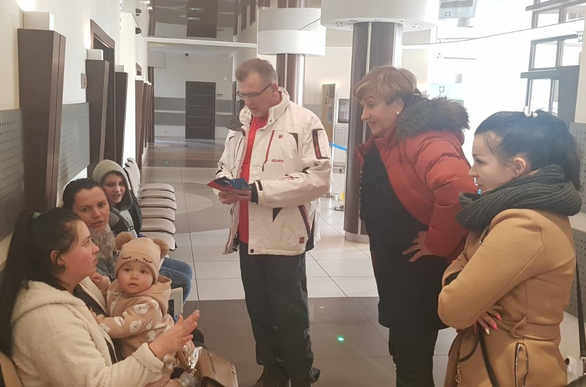 Korytarz urzędu. Z lewej strony, pod ścianą stoją krzesła, na których siedzą dwie kobiety z dziećmi. Z prawej strony na przeciwko siedzących kobiet stoją dwie kobiety i mężczyzna