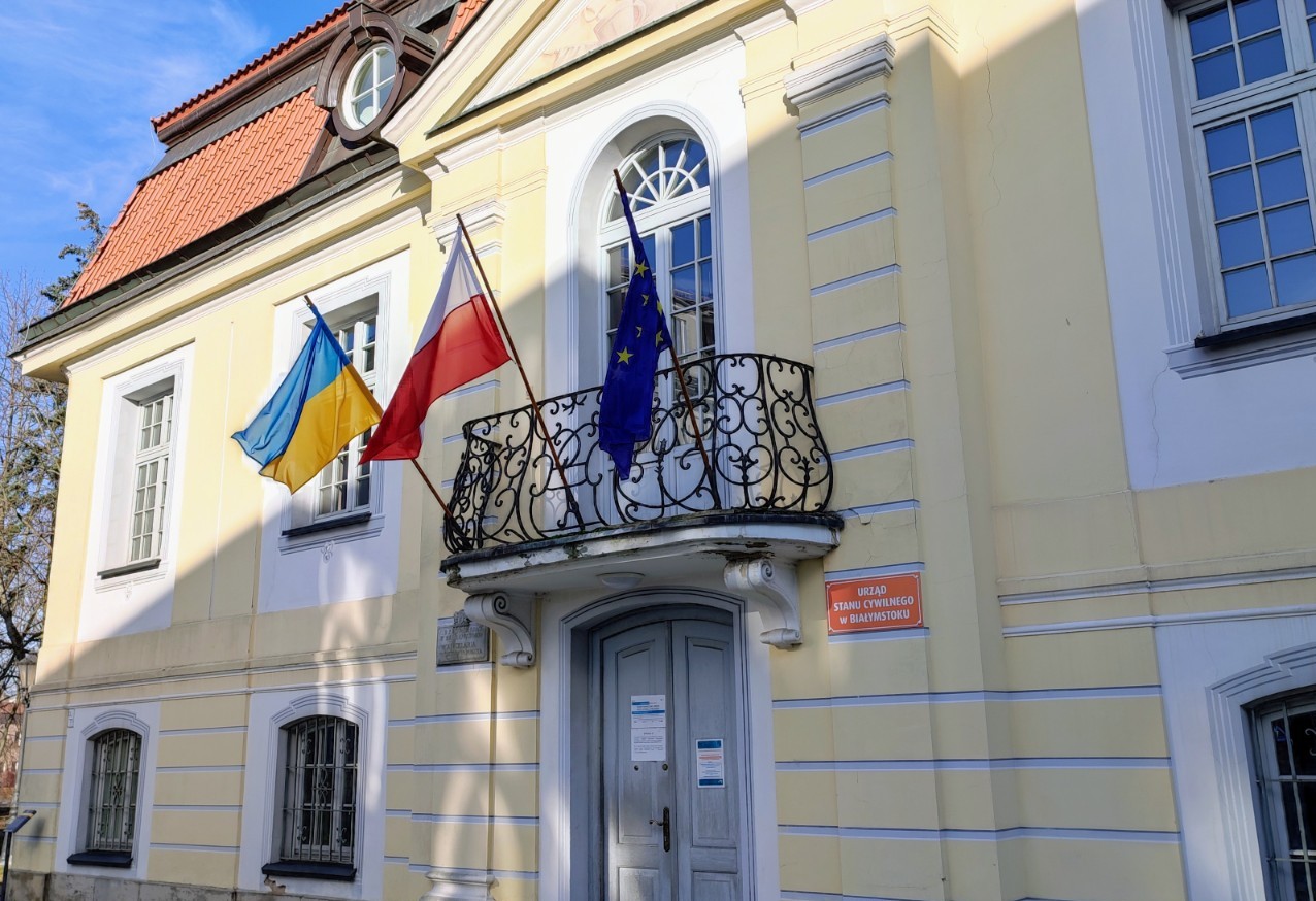 Kremowa fasada Pałacyku Gościnnego w Białymstoku. Z balkonu nad drzwiami powiewają trzy flagi; od lewej Ukrainy (niebiesko-żółta) polska (biało-czerwona) i unijna (granatowa , pośrodku z okręgiem z gwiazdek)