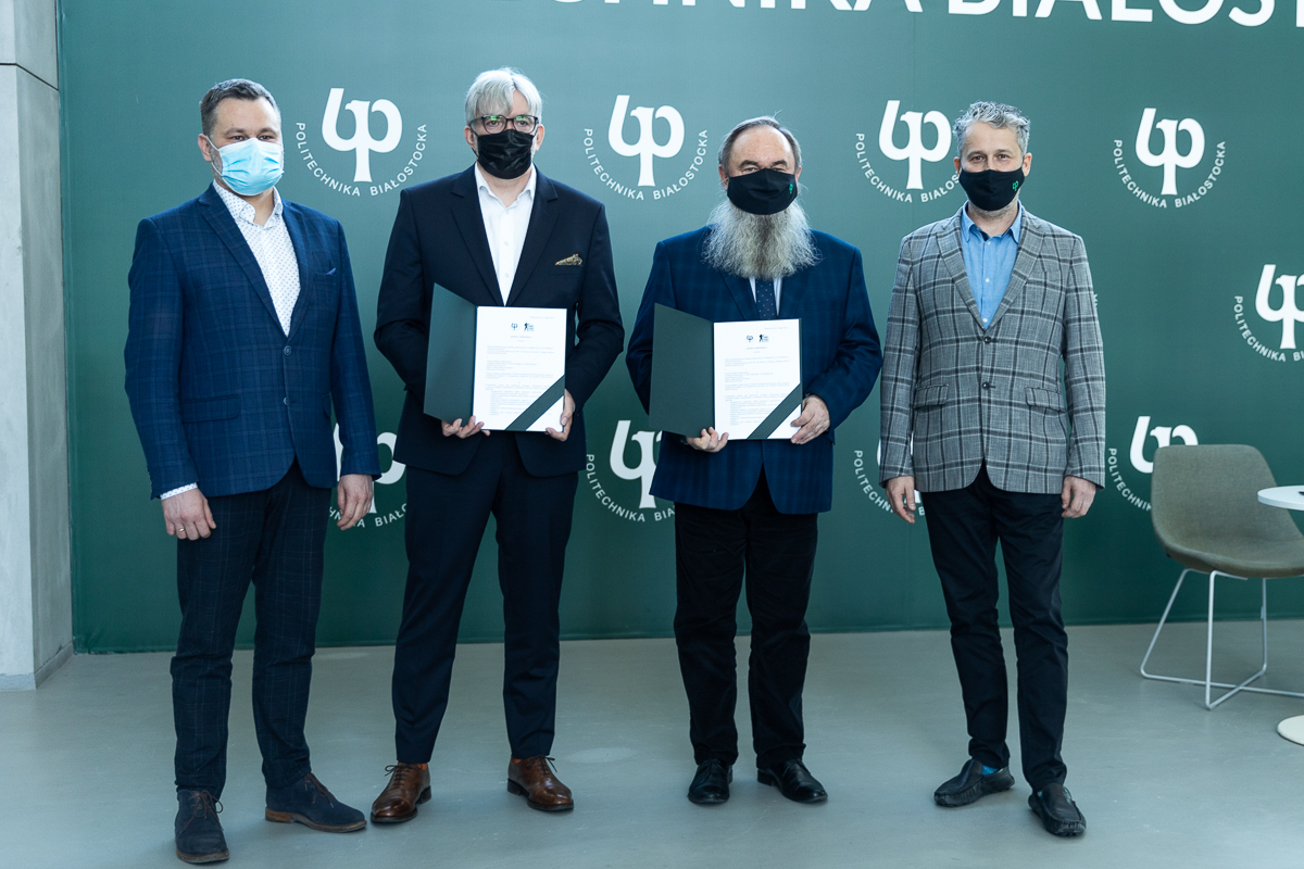 Na tle zielonej ścianki w znaki Politechniki Białostockiej stoi czterech mężczyzn, dwóch stojących w środku trzyma przed sobą podpisane umowy